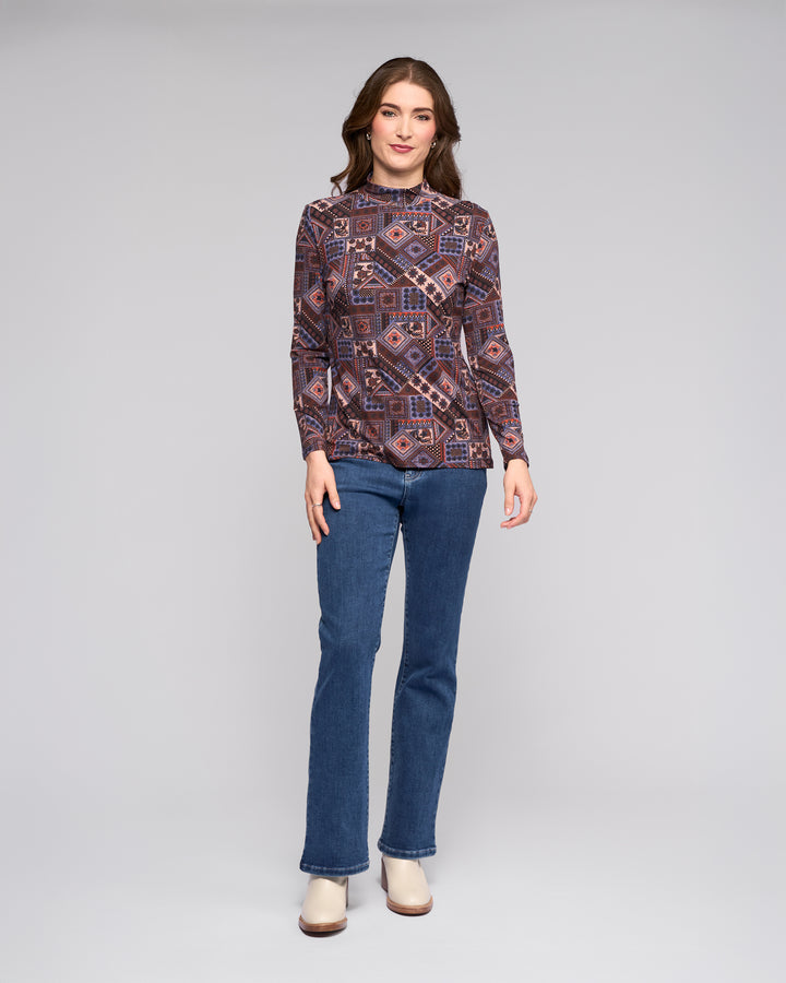 A woman poses in a studio wearing the Vassalli Foundation Top - Rue, styled with blue jeans and beige ankle boots, set against a plain light gray background.