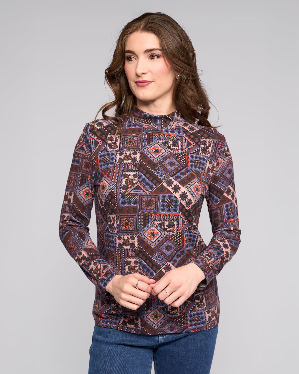 A woman with long brown hair wears the Vassalli Foundation Top - Rue, a long-sleeve high-neck style in blue, purple, and pink, paired with blue jeans, standing against a plain light gray background.
