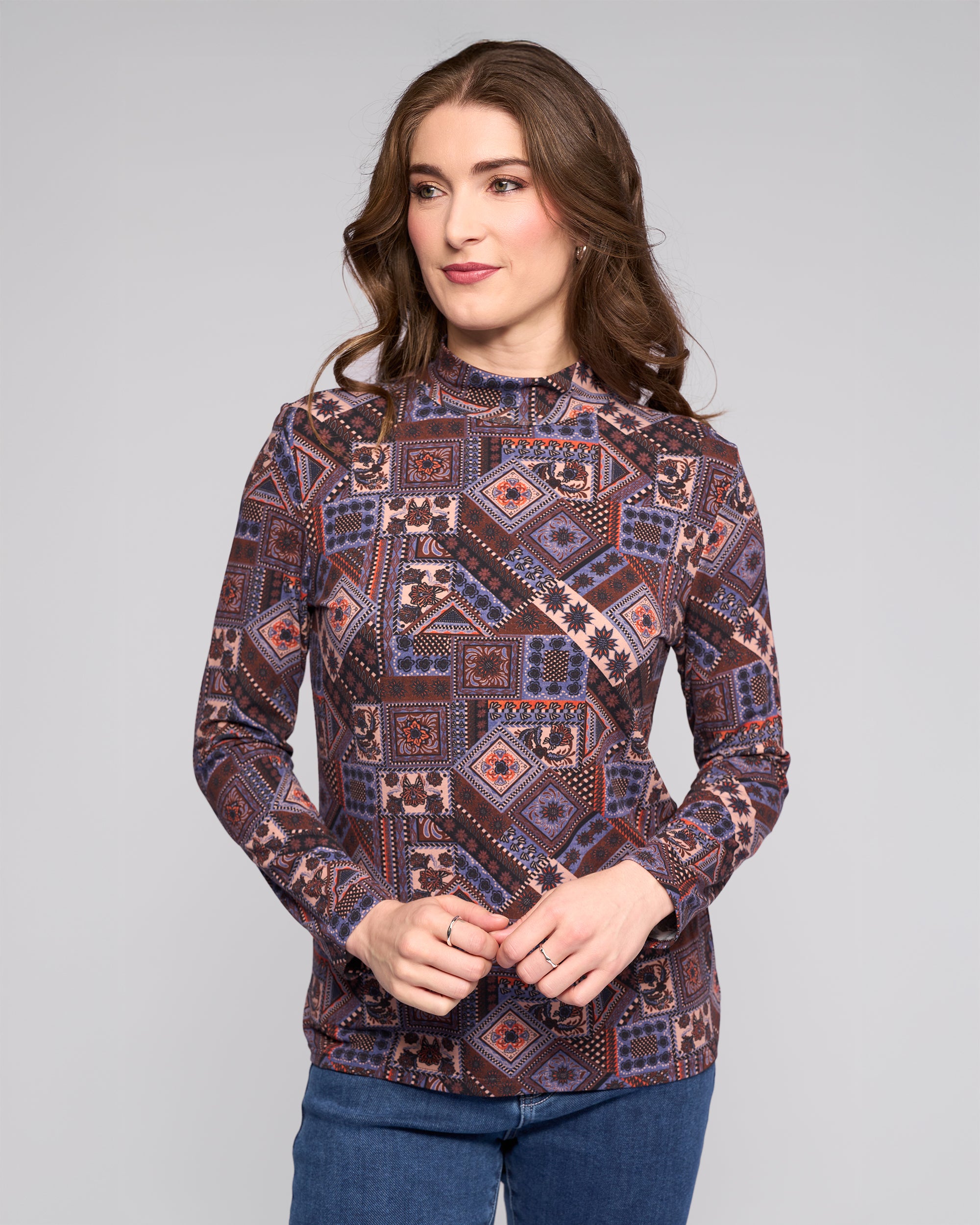 A woman with long brown hair wears the Vassalli Foundation Top - Rue, a long-sleeve high-neck style in blue, purple, and pink, paired with blue jeans, standing against a plain light gray background.