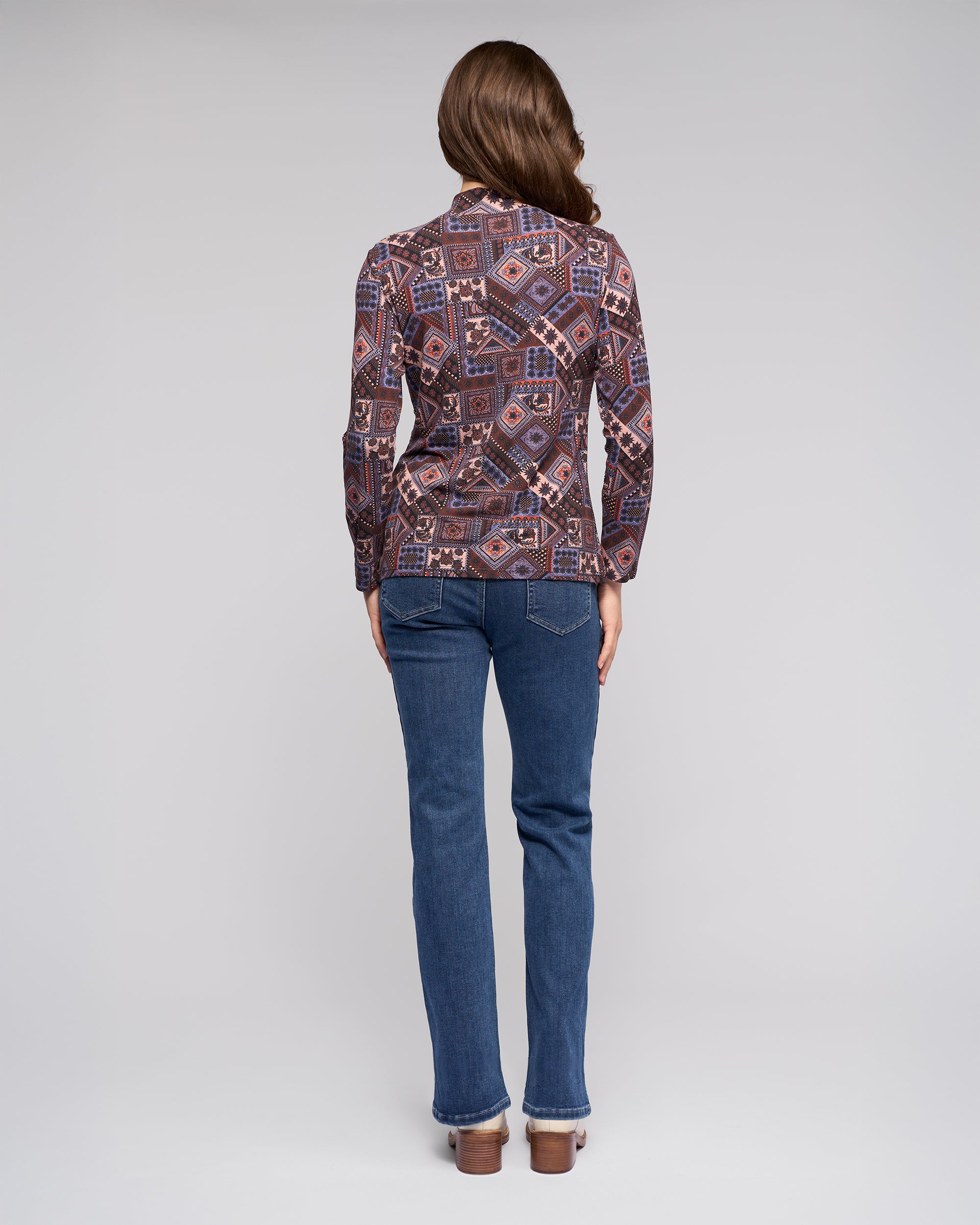 A person with long brown hair stands facing away, wearing the Vassalli Foundation Top - Rue and blue jeans against a plain light gray background.