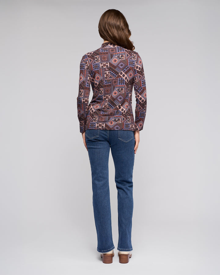 A person with long brown hair stands facing away, wearing the Vassalli Foundation Top - Rue and blue jeans against a plain light gray background.