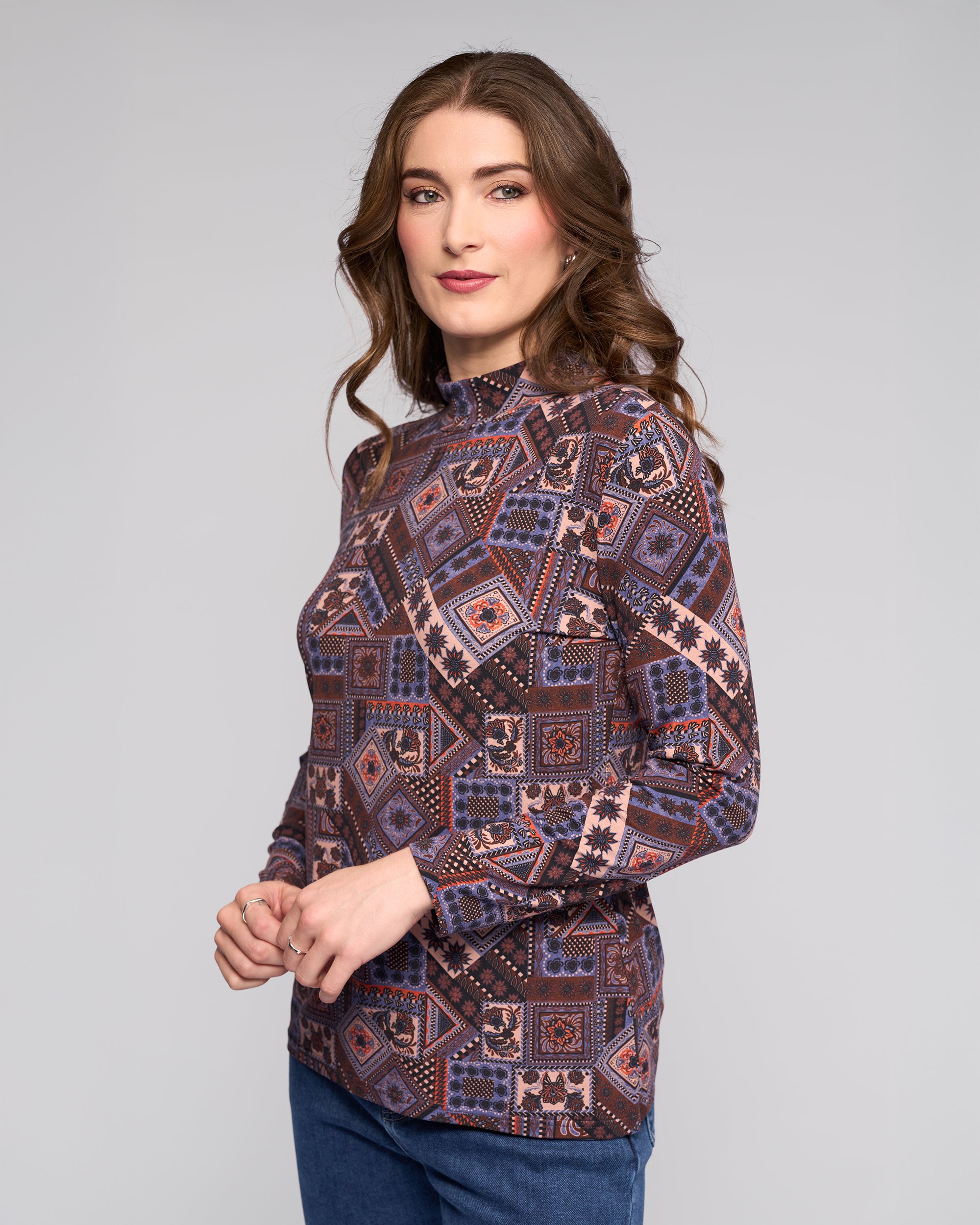 A woman with wavy brown hair stands sideways, wearing the Vassalli Foundation Top - Rue featuring blue, purple, and red geometric shapes. She looks at the camera against a plain gray background.