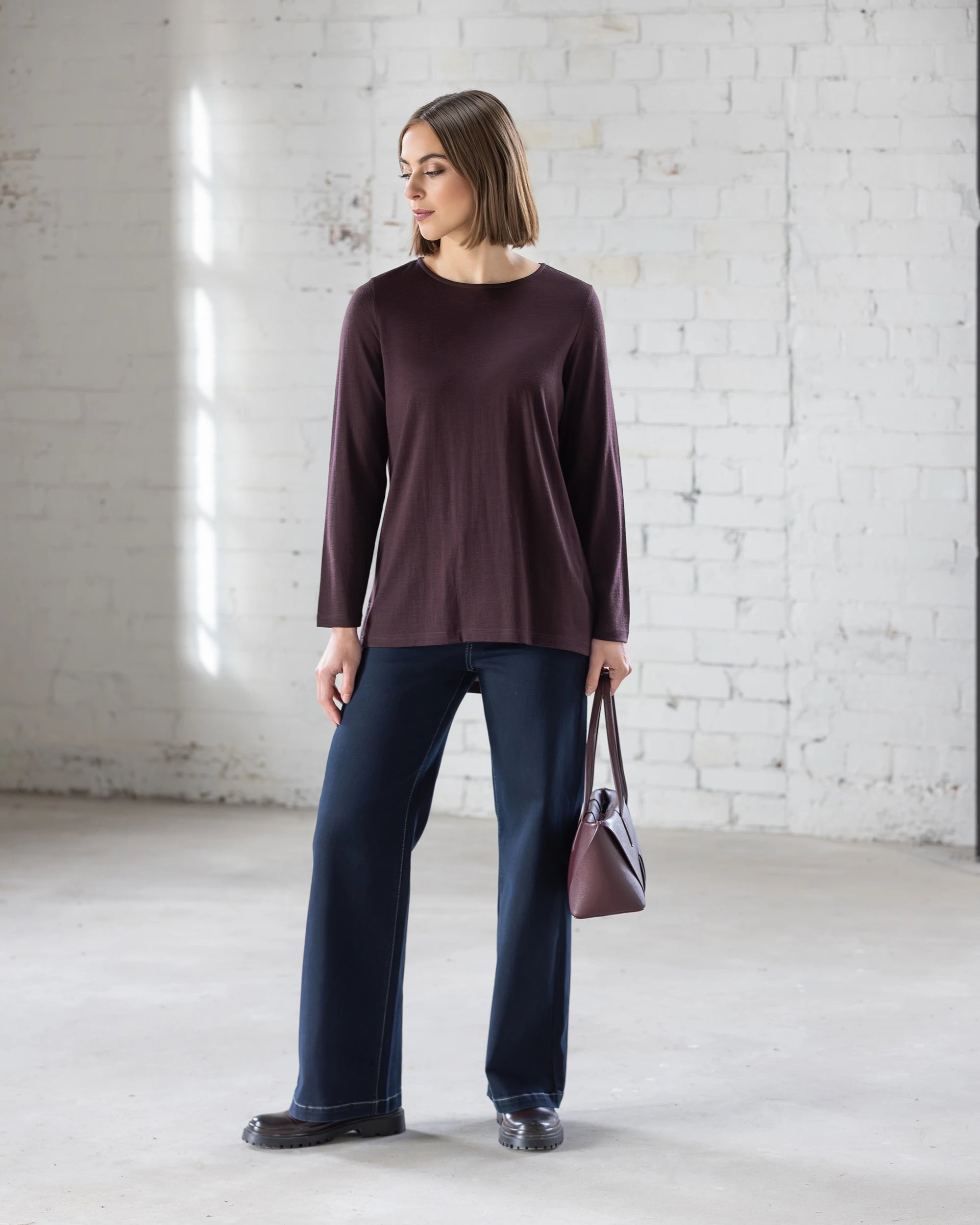 A woman stands in a bright, minimal room with white brick walls, wearing the Vassalli Ivey Merino Top in Berry, wide-leg dark jeans, and black shoes. She holds a brown handbag and gazes to the side.