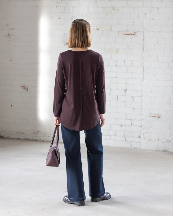 A woman with shoulder-length hair stands indoors facing a white brick wall, wearing the Vassalli Ivey Merino Top in Berry, wide-leg jeans, black shoes, and carrying a matching purple handbag—perfect for chic winter wear.