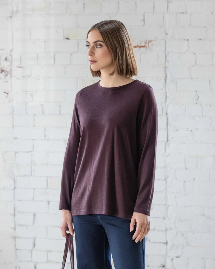 A woman with straight brown hair wears the Vassalli Ivey Merino Top in Berry and dark pants, standing before a white brick wall as she looks left and holds a brown bag—an ideal choice for winter style.