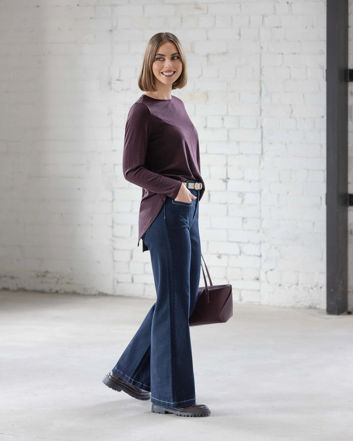 A woman with a bob haircut wears the Vassalli Ivey Merino Top in Berry, dark flared jeans, and black shoes, standing sideways and smiling before a white brick wall while holding a maroon handbag—perfect for winter.