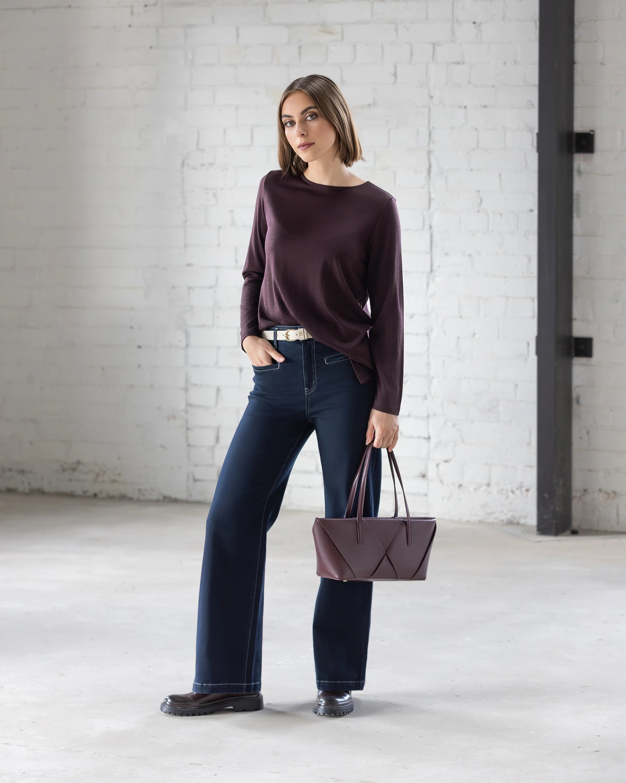 A woman poses indoors against a white brick wall, wearing the Vassalli Ivey Merino Top in Berry with wide-leg blue jeans and black shoes. She holds a matching purple handbag and looks at the camera in a relaxed stance.