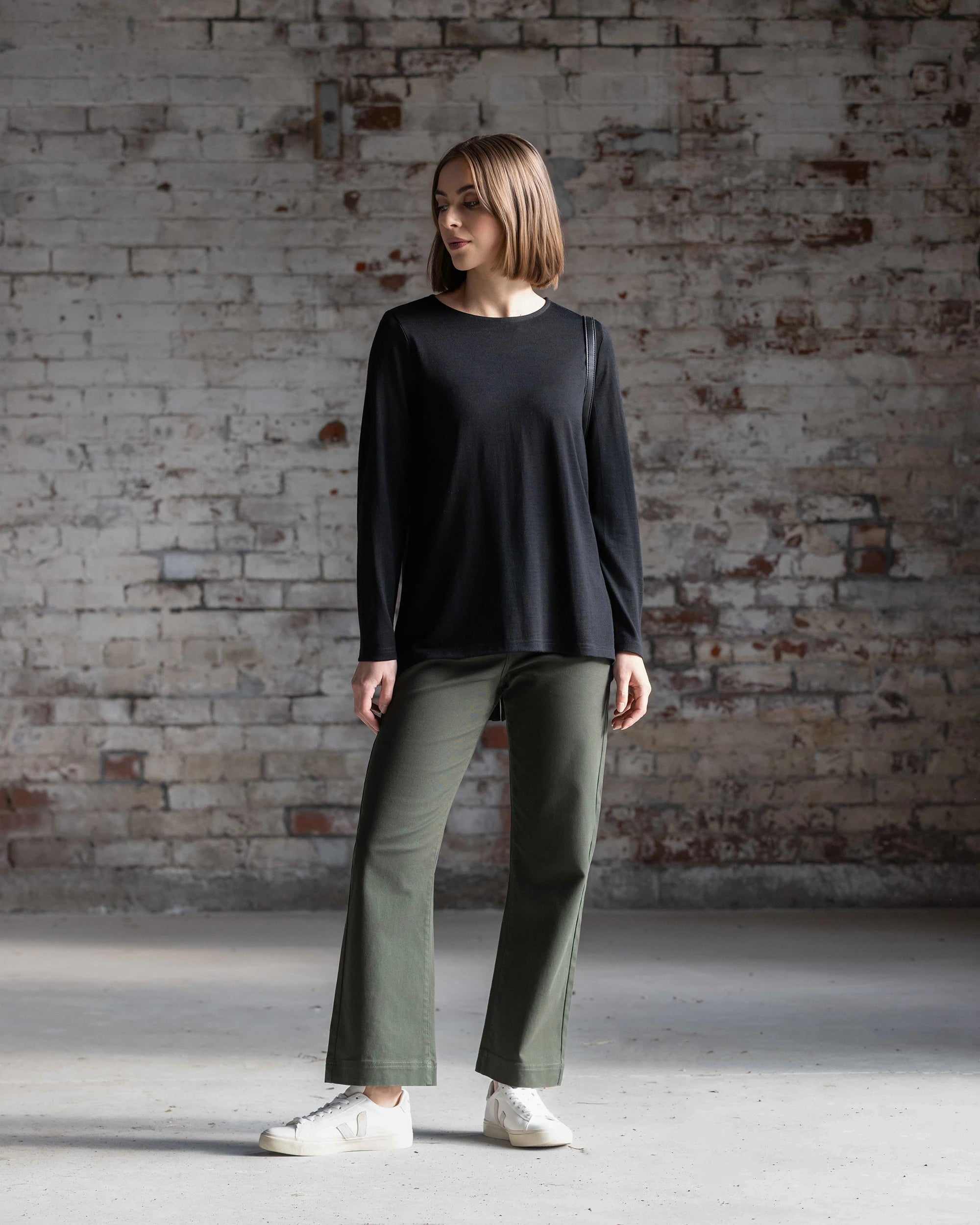 A young woman stands by a weathered brick wall, wearing the Vassalli Ivey Merino Top in black, relaxed olive green pants, and white sneakers. She gazes to the side with a casual and relaxed posture.