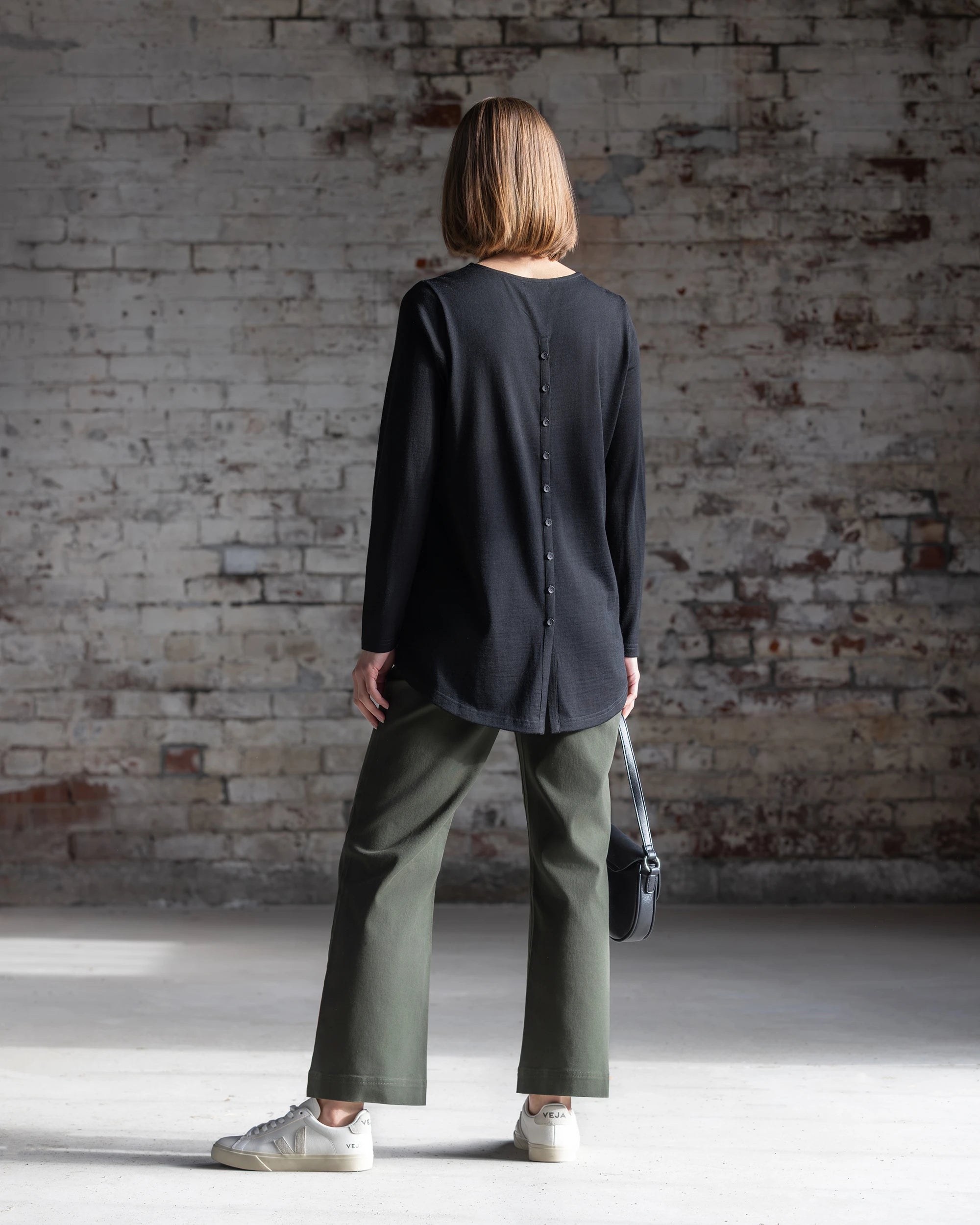 A woman with straight, shoulder-length hair faces a brick wall, wearing the Vassalli Ivey Merino Top in black, olive green pants, white sneakers, and holding a small black handbag.