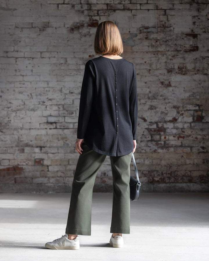 A woman with straight, shoulder-length hair faces a brick wall, wearing the Vassalli Ivey Merino Top in black, olive green pants, white sneakers, and holding a small black handbag.