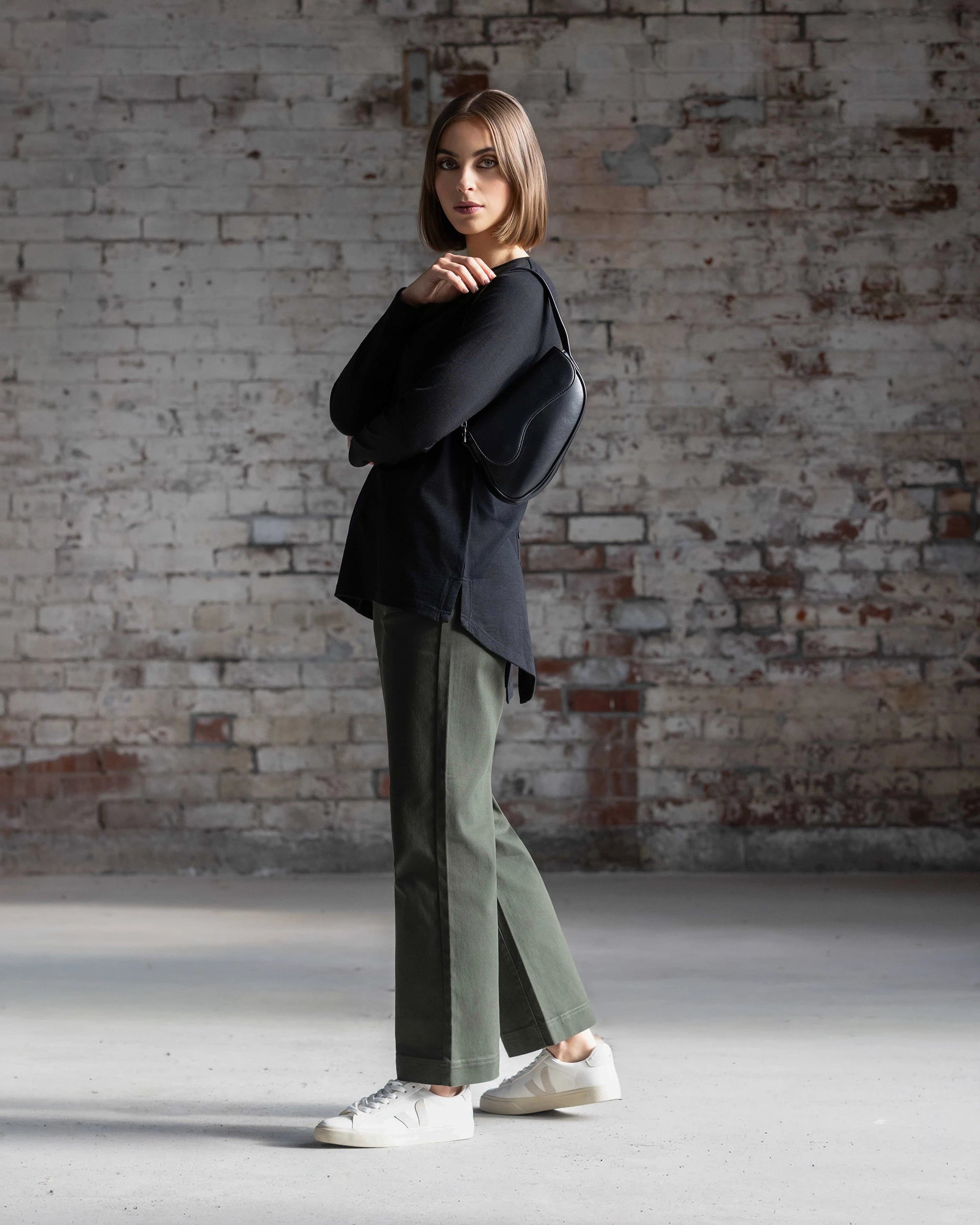 A woman with straight brown hair stands sideways by a brick wall, wearing the Vassalli Ivey Merino Top in black, green pants, white sneakers, and carrying a black shoulder bag.
