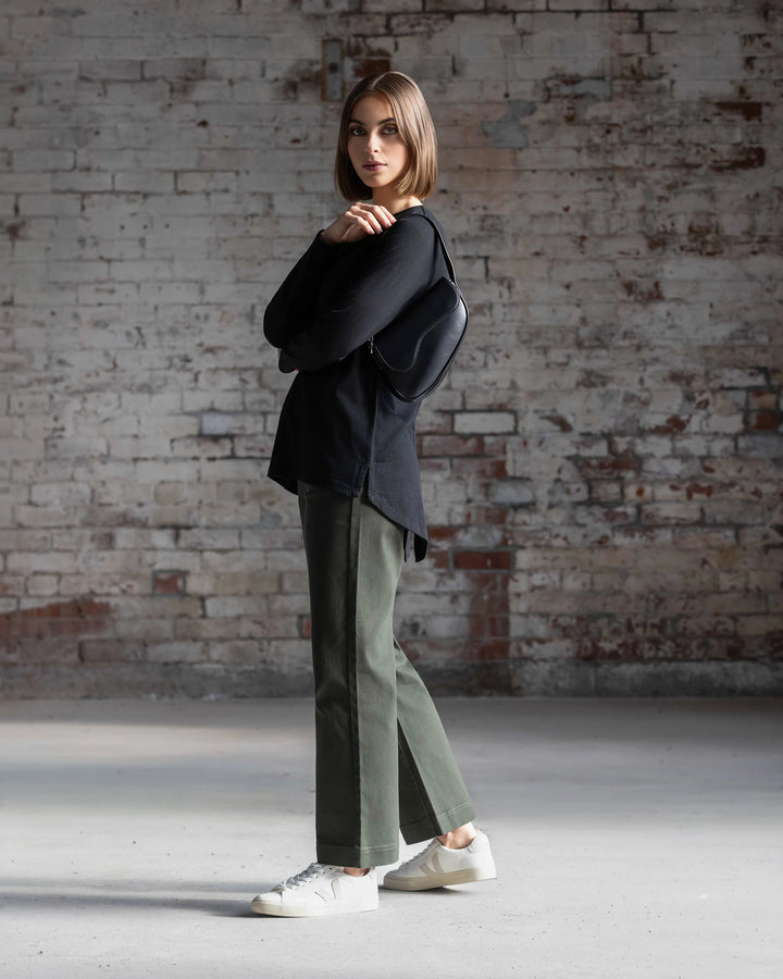 A woman with straight brown hair stands sideways by a brick wall, wearing the Vassalli Ivey Merino Top in black, green pants, white sneakers, and carrying a black shoulder bag.