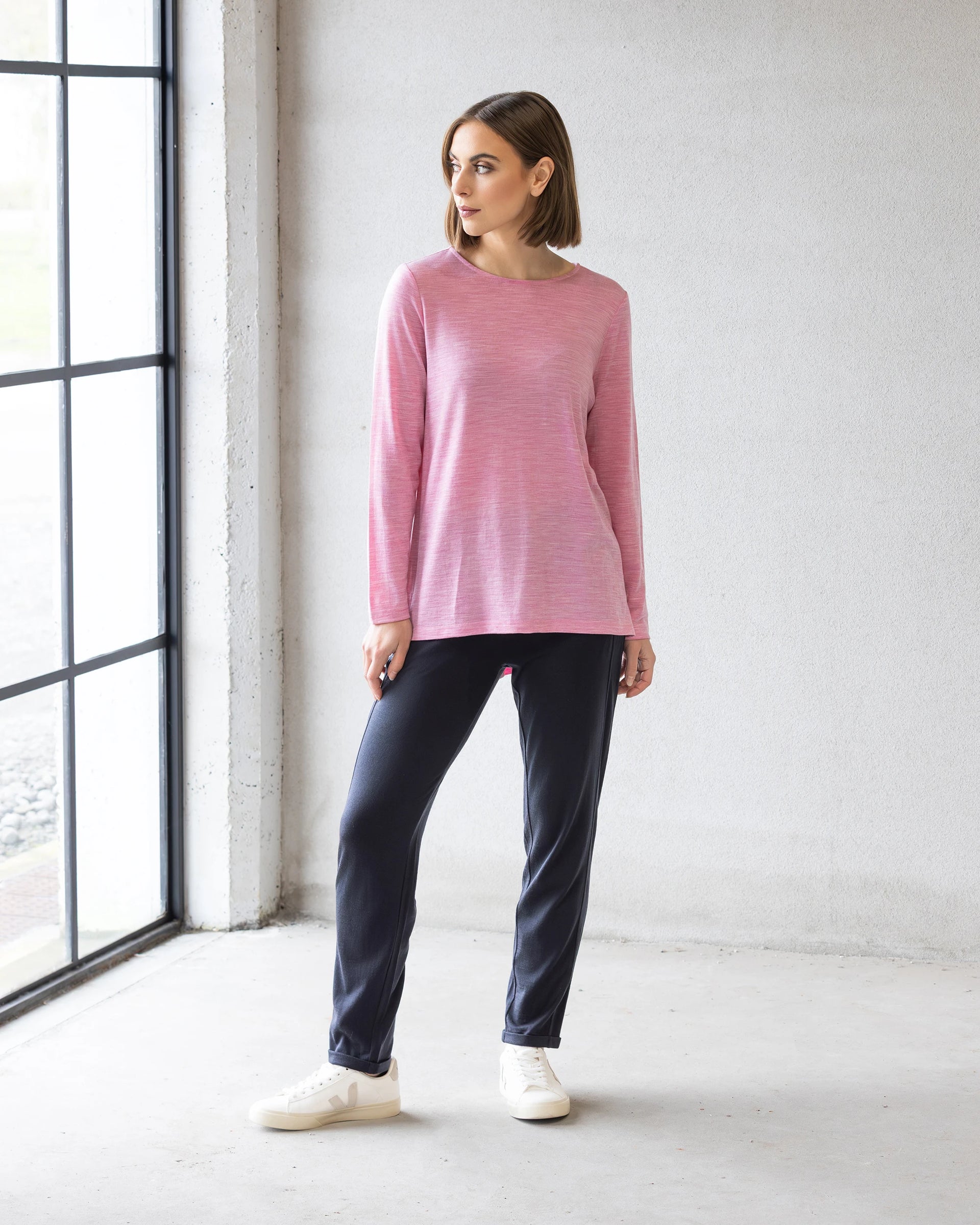 Indoors by a large window, a person wears the Vassalli Ivey Merino Top in Pink Marle with dark pants and white sneakers—perfect for winter layering. The space has light walls and a concrete floor.
