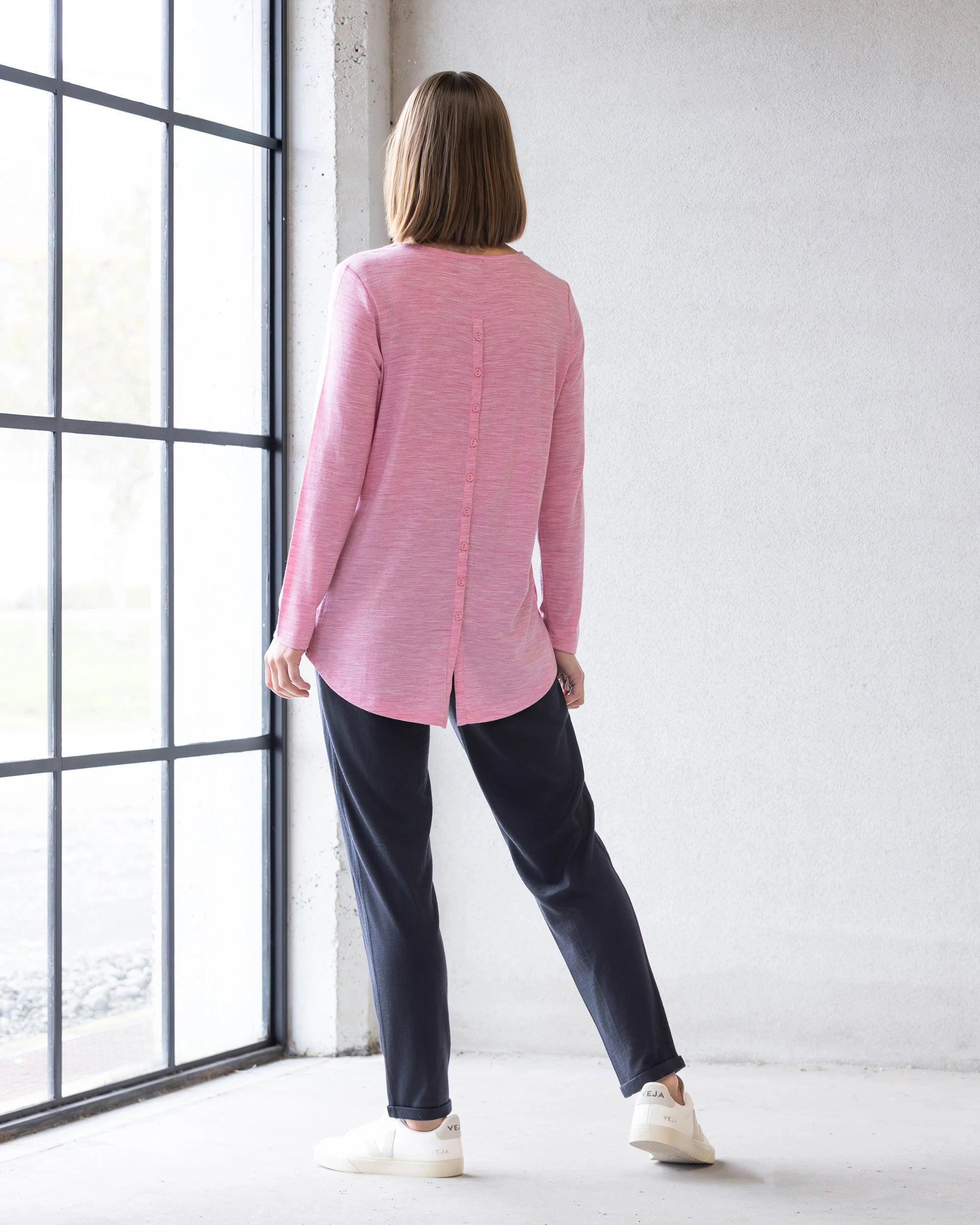 Indoors by a large window, a person with straight, shoulder-length hair wears the Vassalli Ivey Merino Top in Pink Marle and dark pants with white sneakers—perfect for winter layering. The top features a vertical seam and back slit.