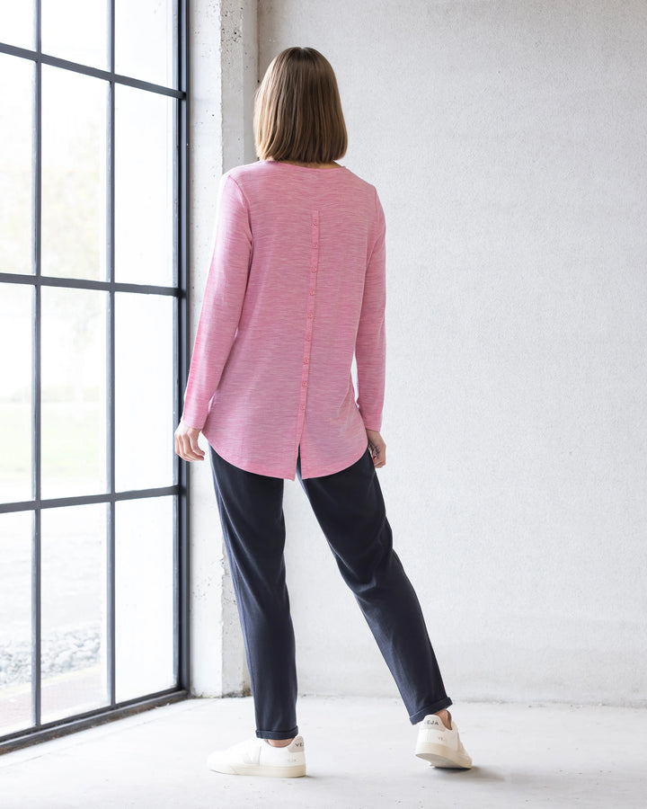 Indoors by a large window, a person with straight, shoulder-length hair wears the Vassalli Ivey Merino Top in Pink Marle and dark pants with white sneakers—perfect for winter layering. The top features a vertical seam and back slit.