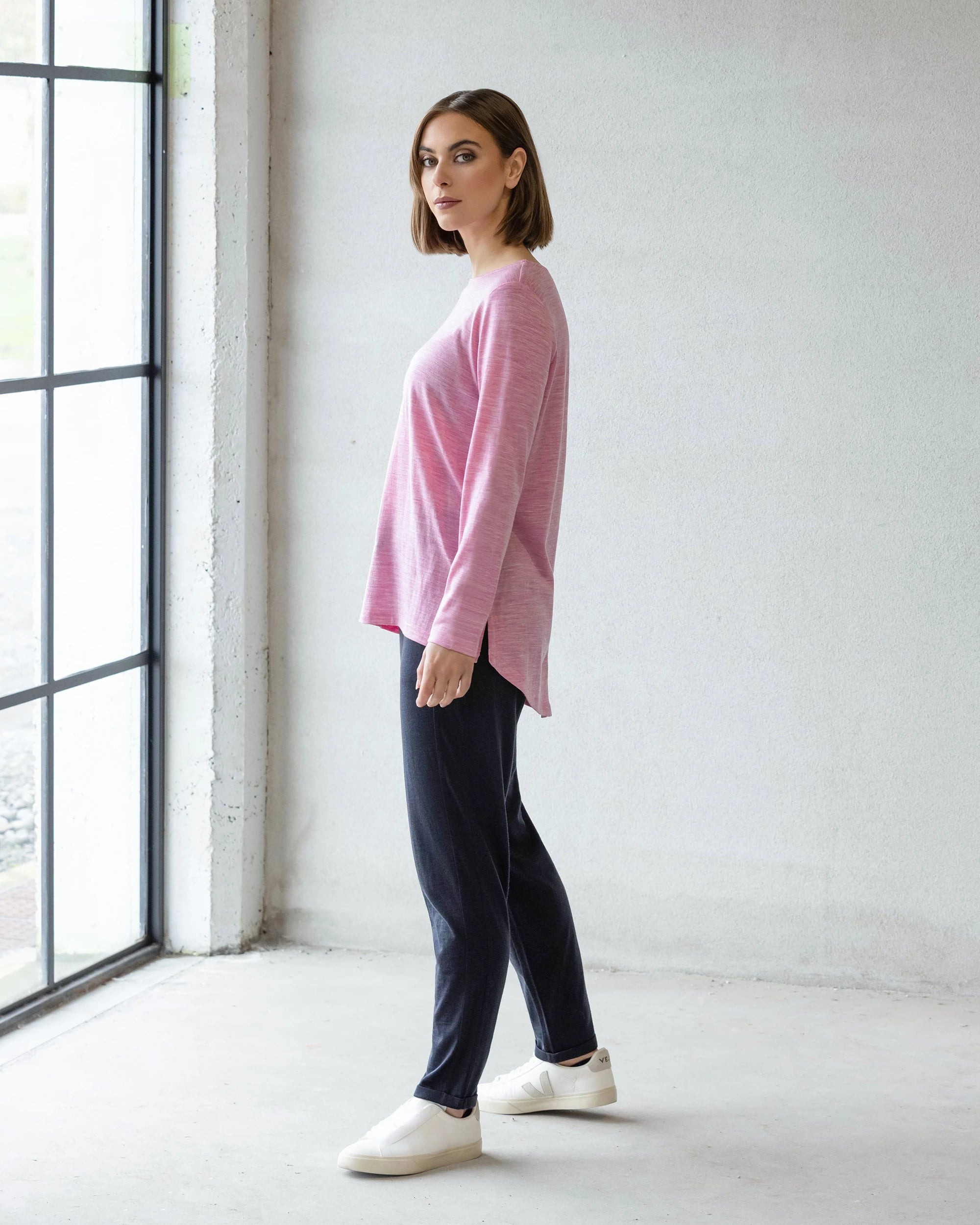 A woman stands indoors near a large window, wearing the Vassalli Ivey Merino Top in Pink Marle, paired with dark pants and white slip-on shoes. She faces forward against a light wall—an ideal look for effortless winter layering.