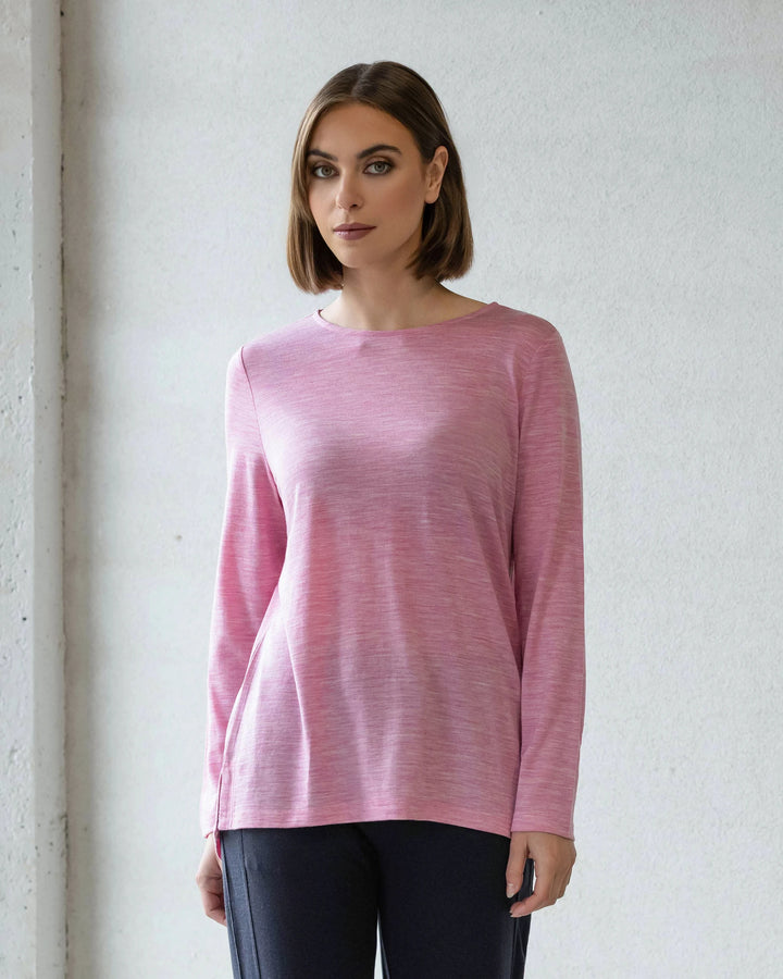 A woman with straight, shoulder-length brown hair wears the Vassalli Ivey Merino Top in Pink Marle and dark pants, standing against a light wall and looking at the camera—an ideal winter layering piece.