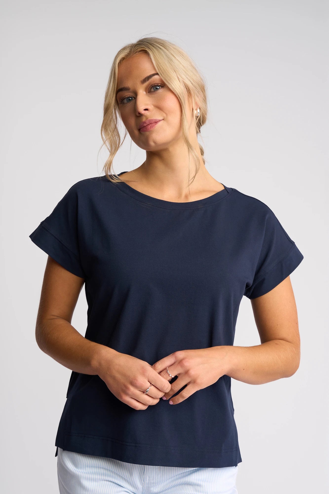 A woman with blonde hair is wearing the Vassalli Thea Top in Ink, featuring a boat neckline, paired with light-colored pants. She stands against a plain light background, looking at the camera with a slight smile.