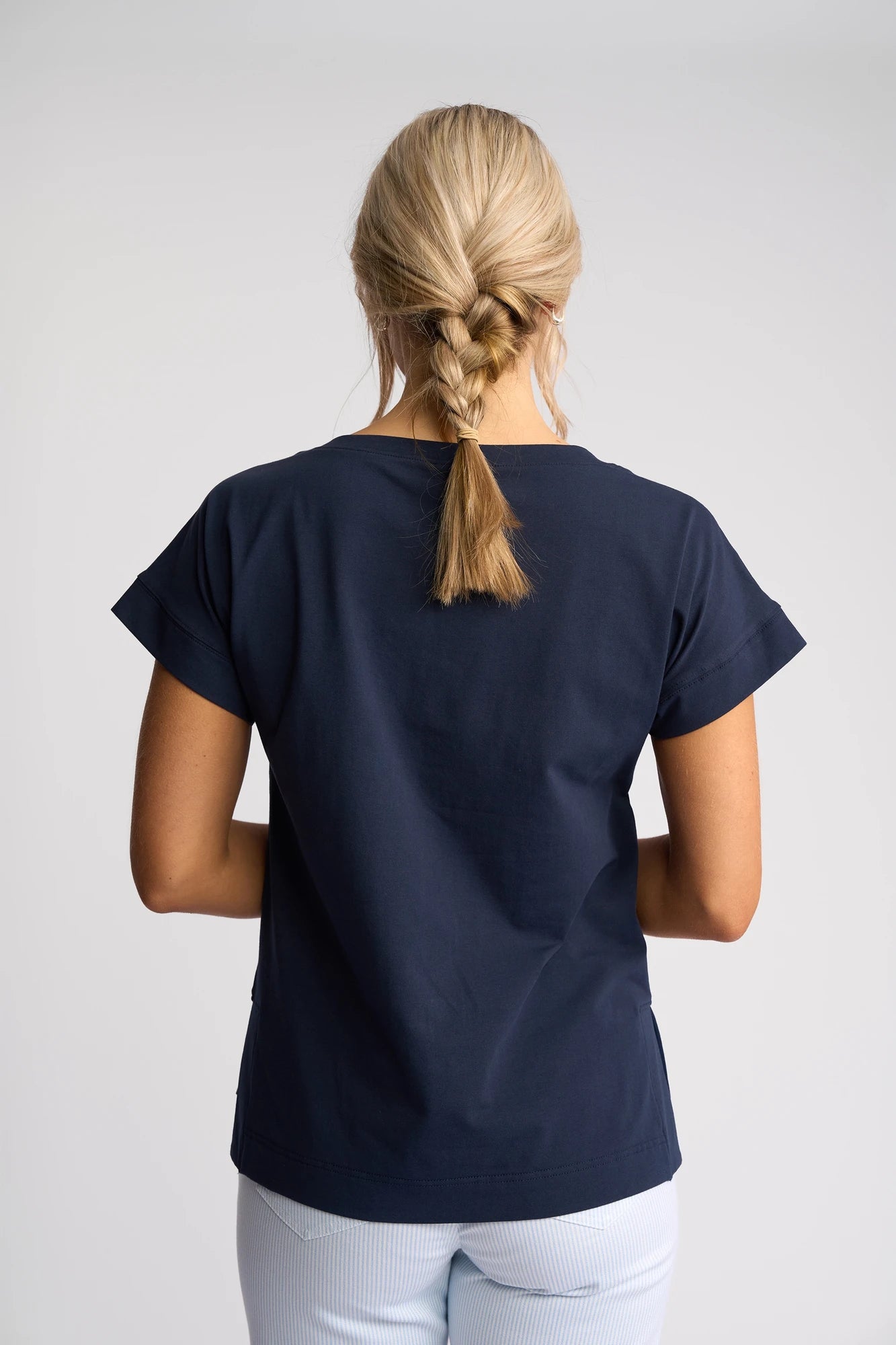 A woman with blonde hair in a braid is standing with her back to the camera, wearing a Vassalli Thea Top in Ink with a boat neckline and light pants against a plain light background.
