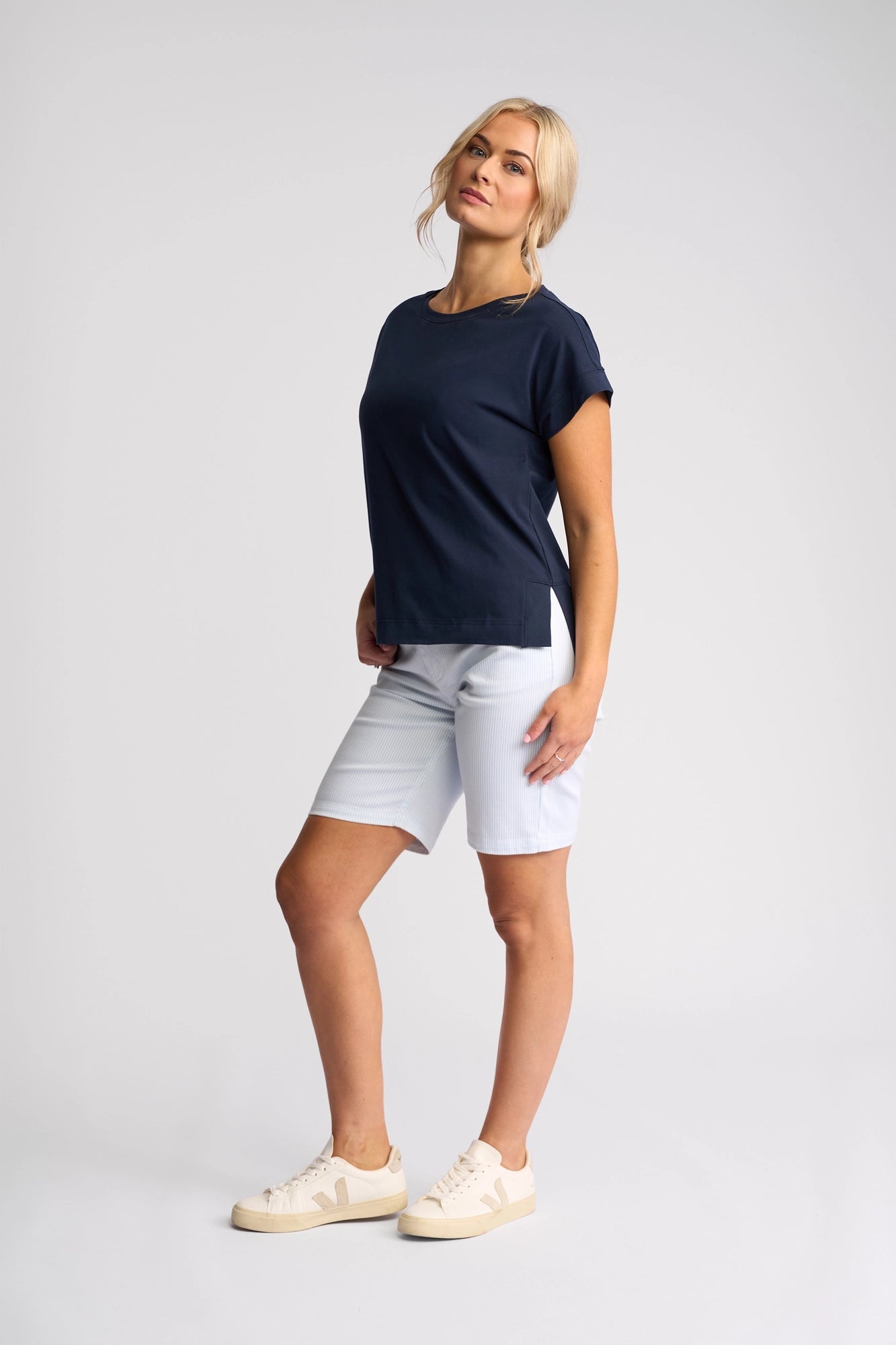 A woman stands against a plain background, wearing the Vassalli Thea Top in Ink, paired with light blue knee-length shorts and cream sneakers. Her blonde hair is styled in a loose updo, and she poses with one leg slightly bent.