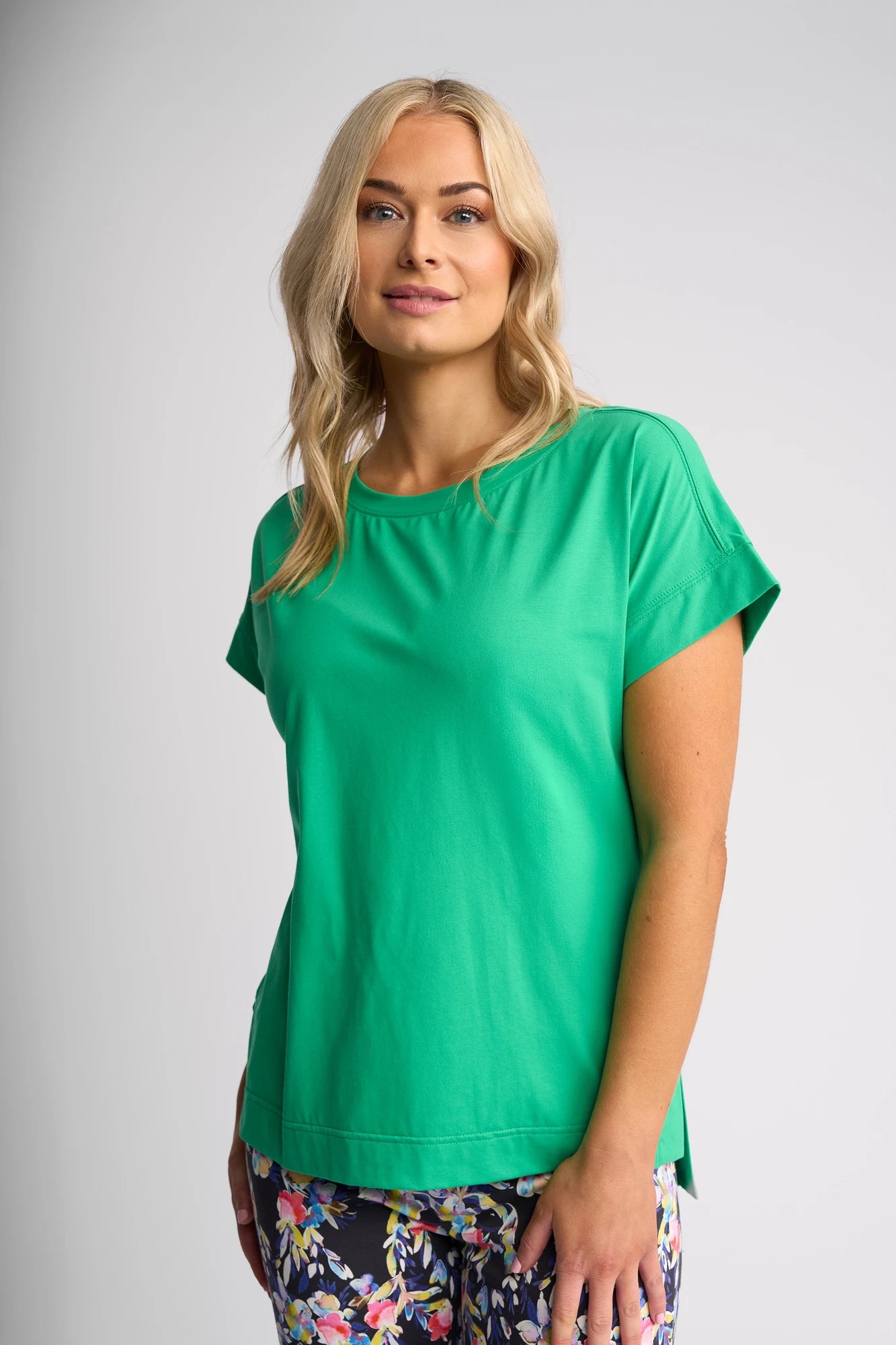 A woman with long blonde hair wears the Vassalli Thea Top in Summer Green and dark floral pants, standing against a plain light gray background while smiling softly at the camera.