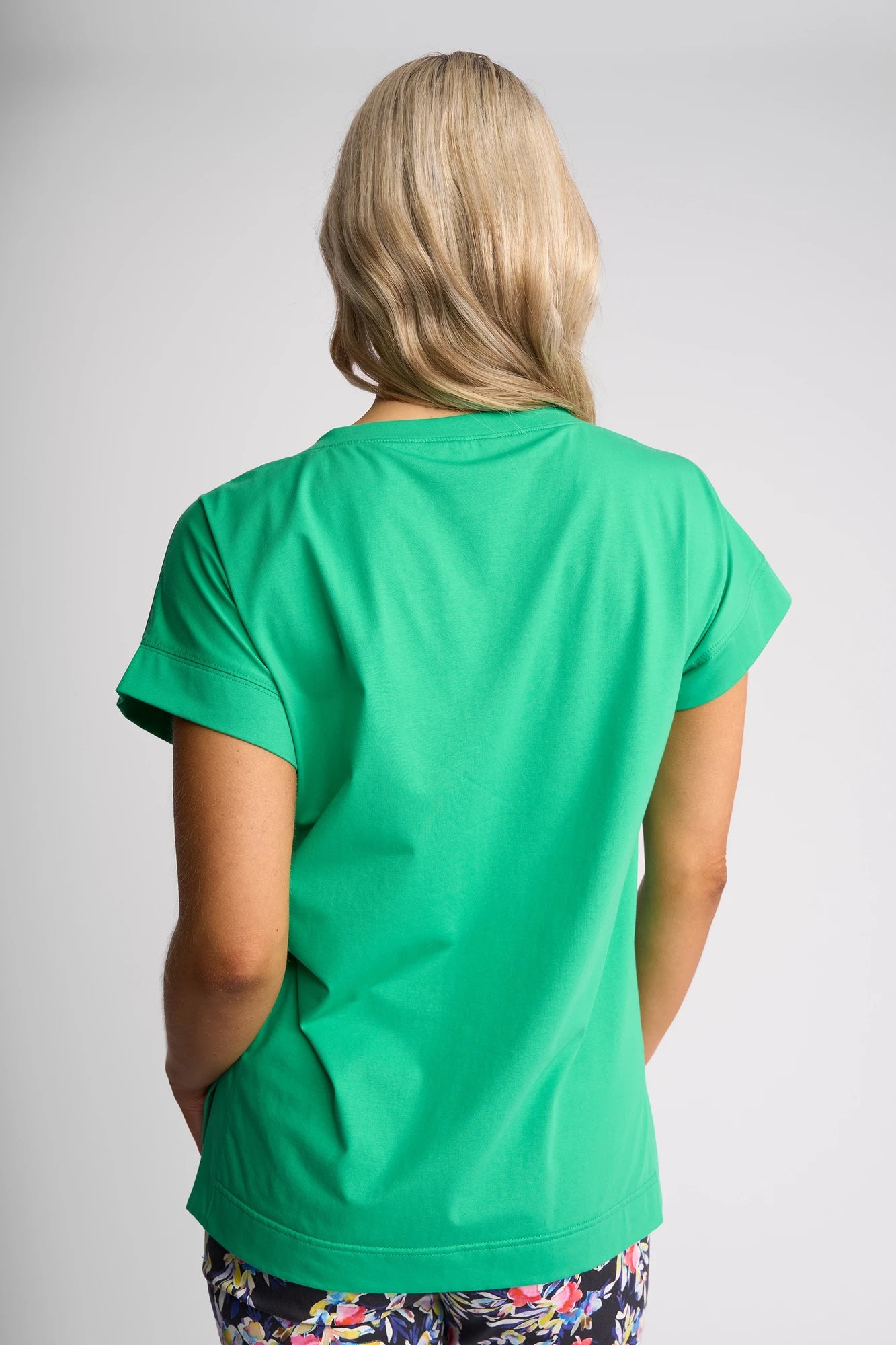 A person with blonde hair faces away, wearing the Vassalli Thea Top in Summer Green—a vibrant, comfortable women’s stretch cotton tee—paired with colorful floral-patterned pants against a plain light gray background.