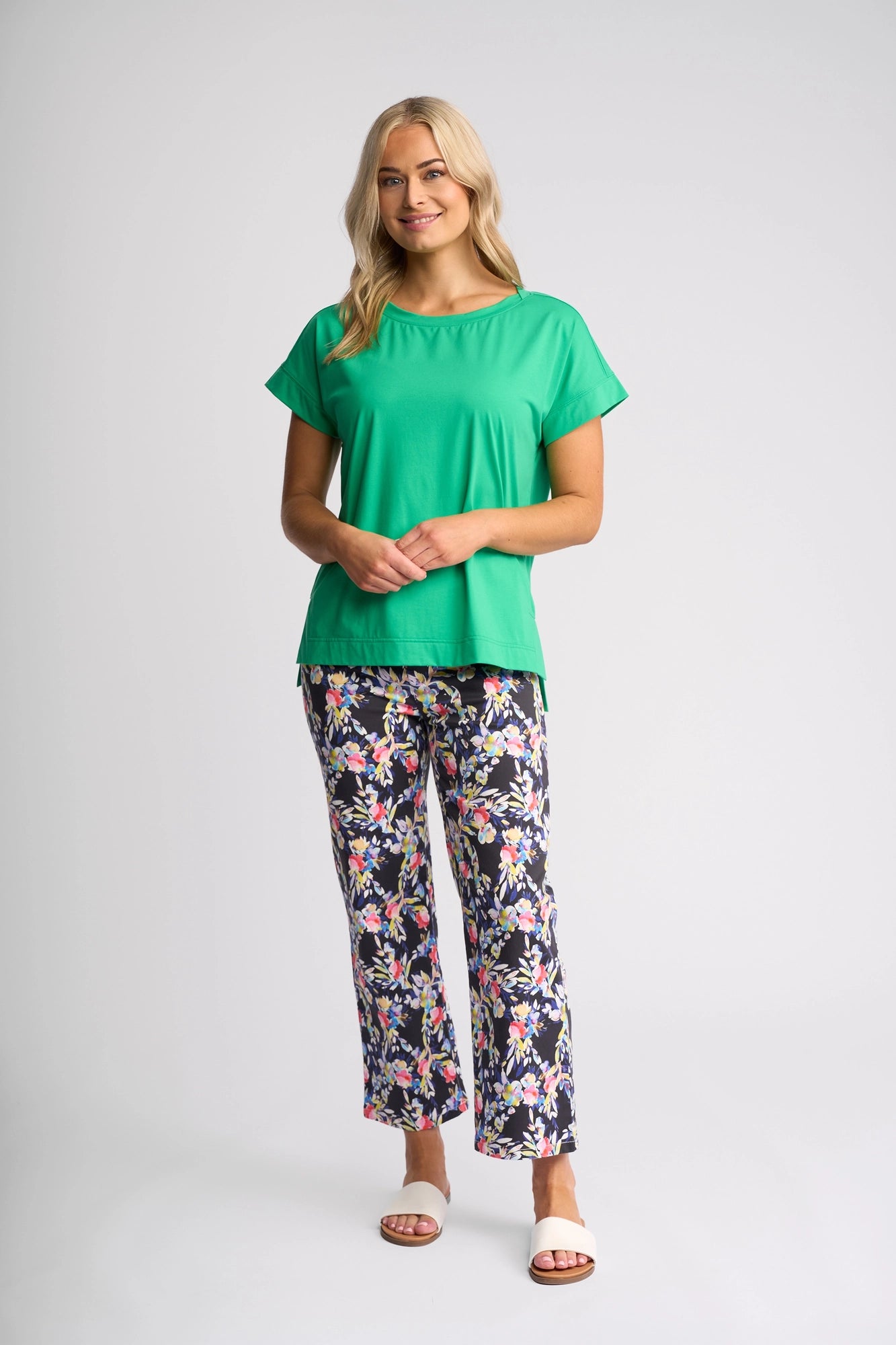 A woman with long blonde hair wears the Vassalli Thea Top in Summer Green, a fashionable cotton tee, paired with floral print pants and white sandals. She stands smiling against a plain light gray background.
