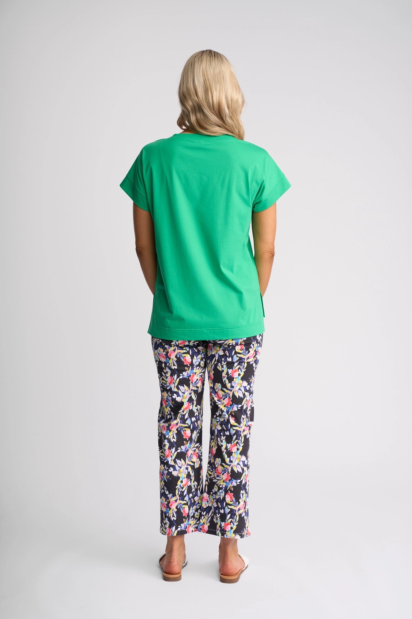 A person stands facing away, wearing the Vassalli Thea Top in Summer Green paired with colorful floral pants and white sandals. The background is plain and light.