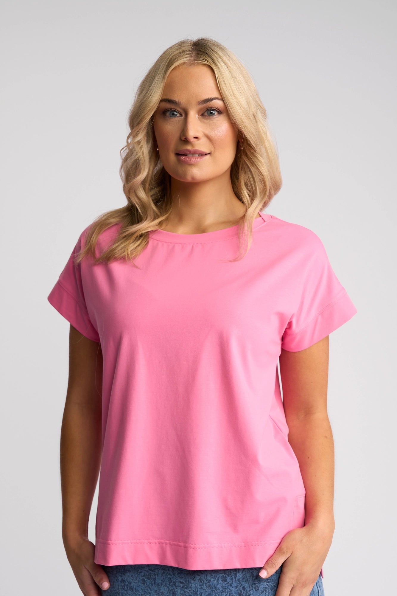 A woman with long blonde hair wears the Vassalli Thea Top in Sweet Pink and blue jeans, standing against a plain light gray background and looking at the camera with a slight smile.