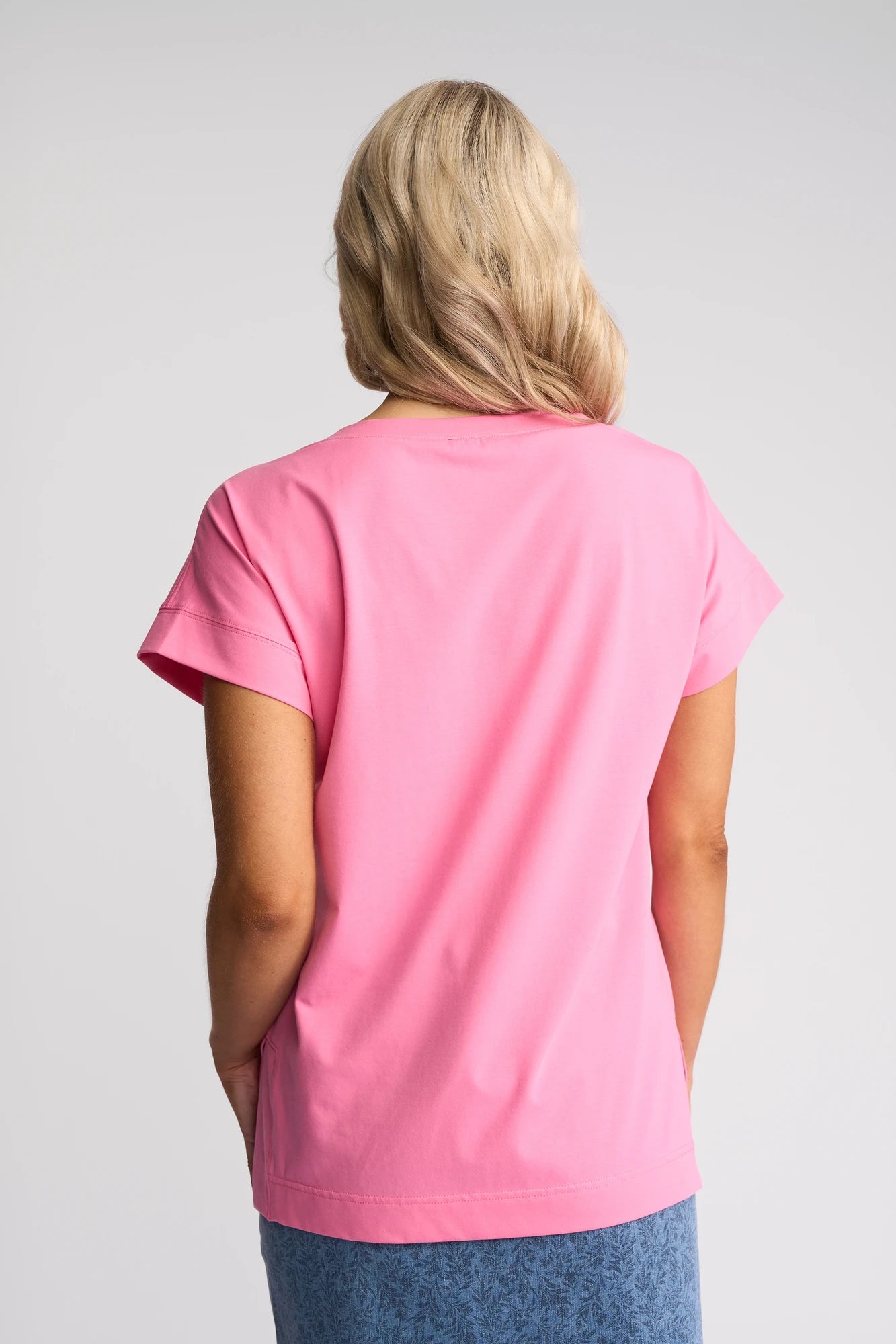 Facing away from the camera, a blonde-haired individual is wearing the Vassalli Thea Top - Sweet Pink, complemented by a blue patterned skirt, with both set against a simple light background.