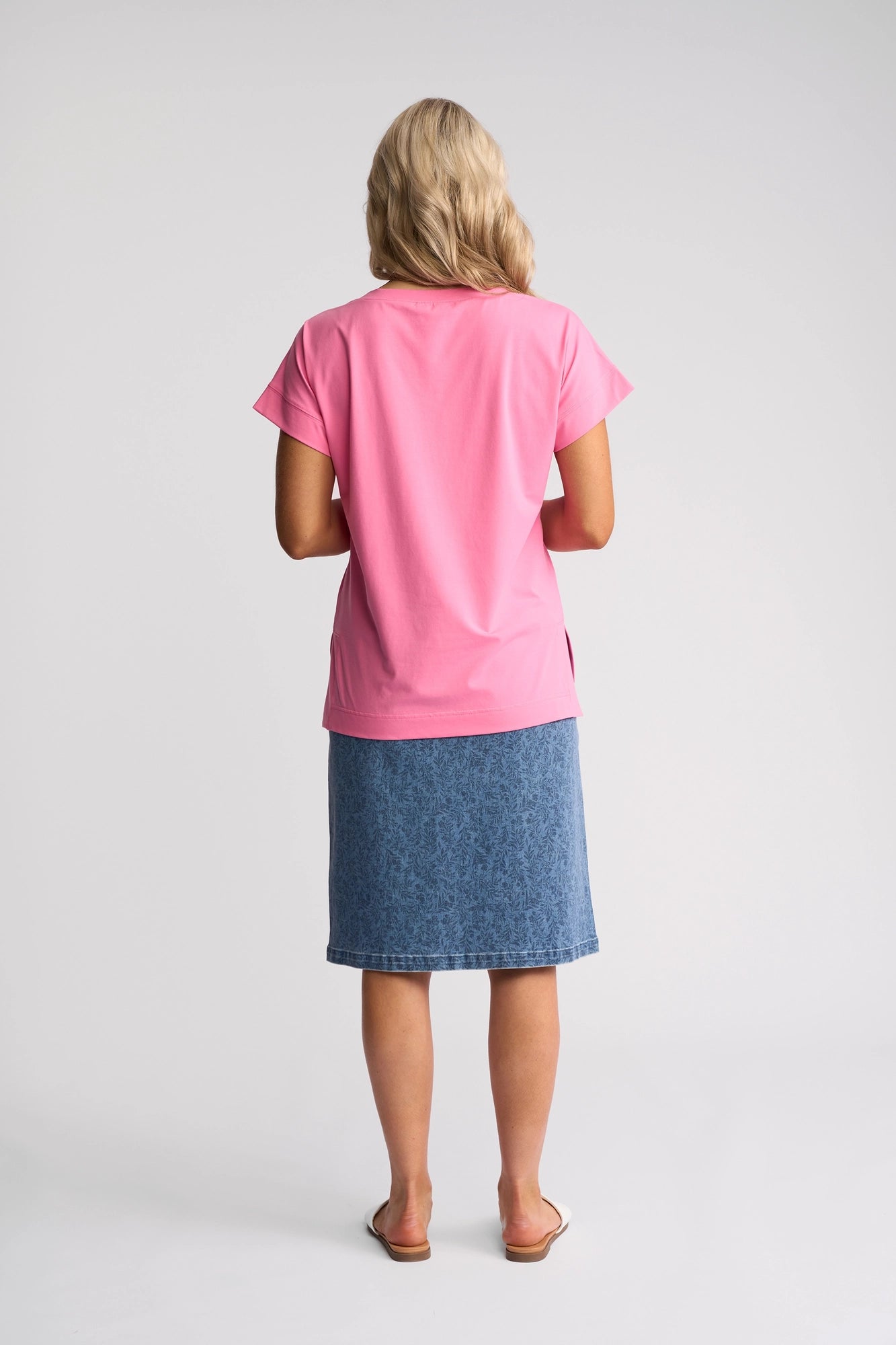 A person with blonde hair stands facing away, wearing the Vassalli Thea Top in Sweet Pink—a soft cotton tee—paired with a blue knee-length denim skirt and white sandals, set against a plain white background.