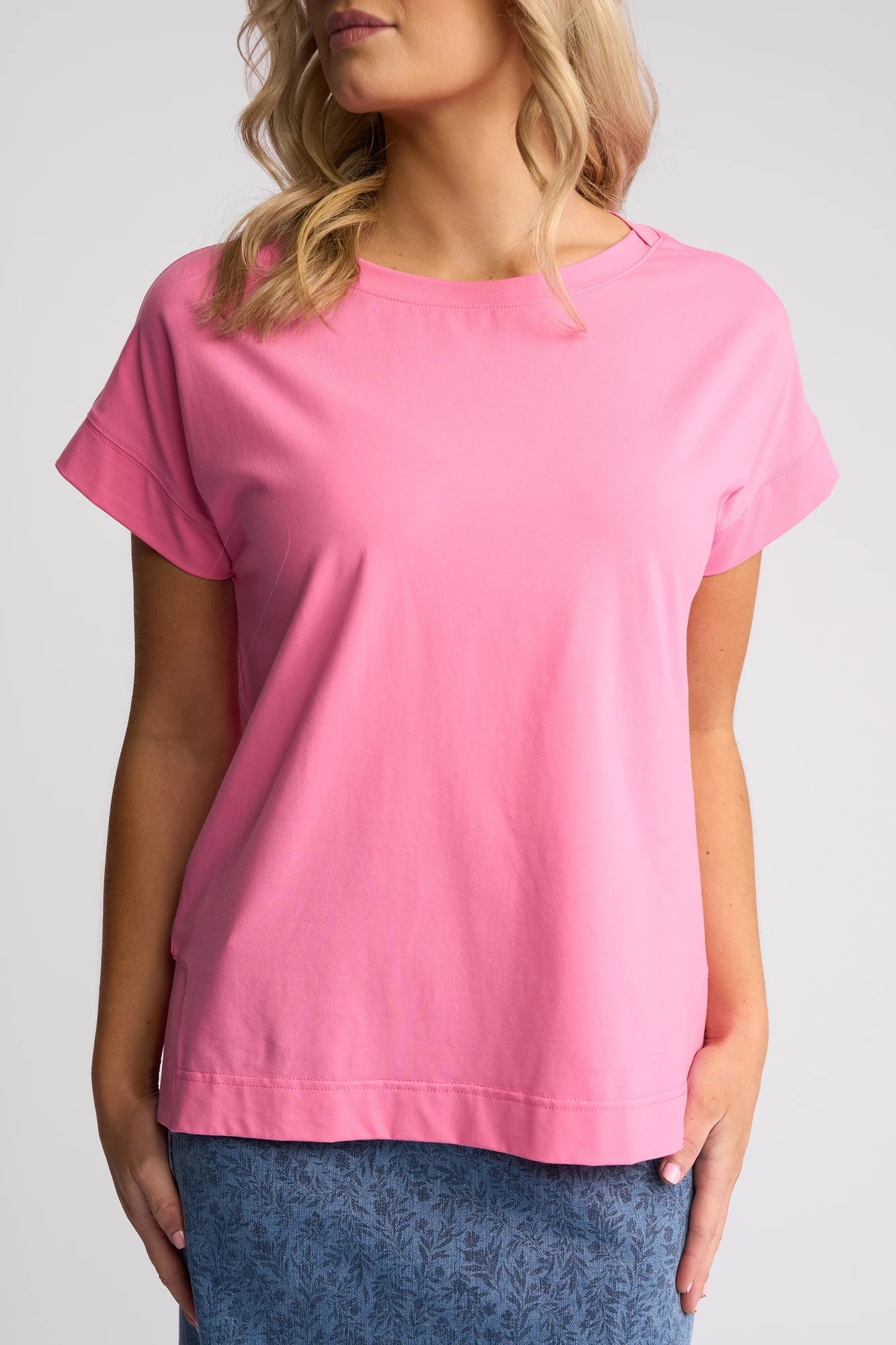 A woman in the Vassalli Thea Top in Sweet Pink, paired with a blue skirt featuring a subtle leafy pattern, stands against a plain light background.