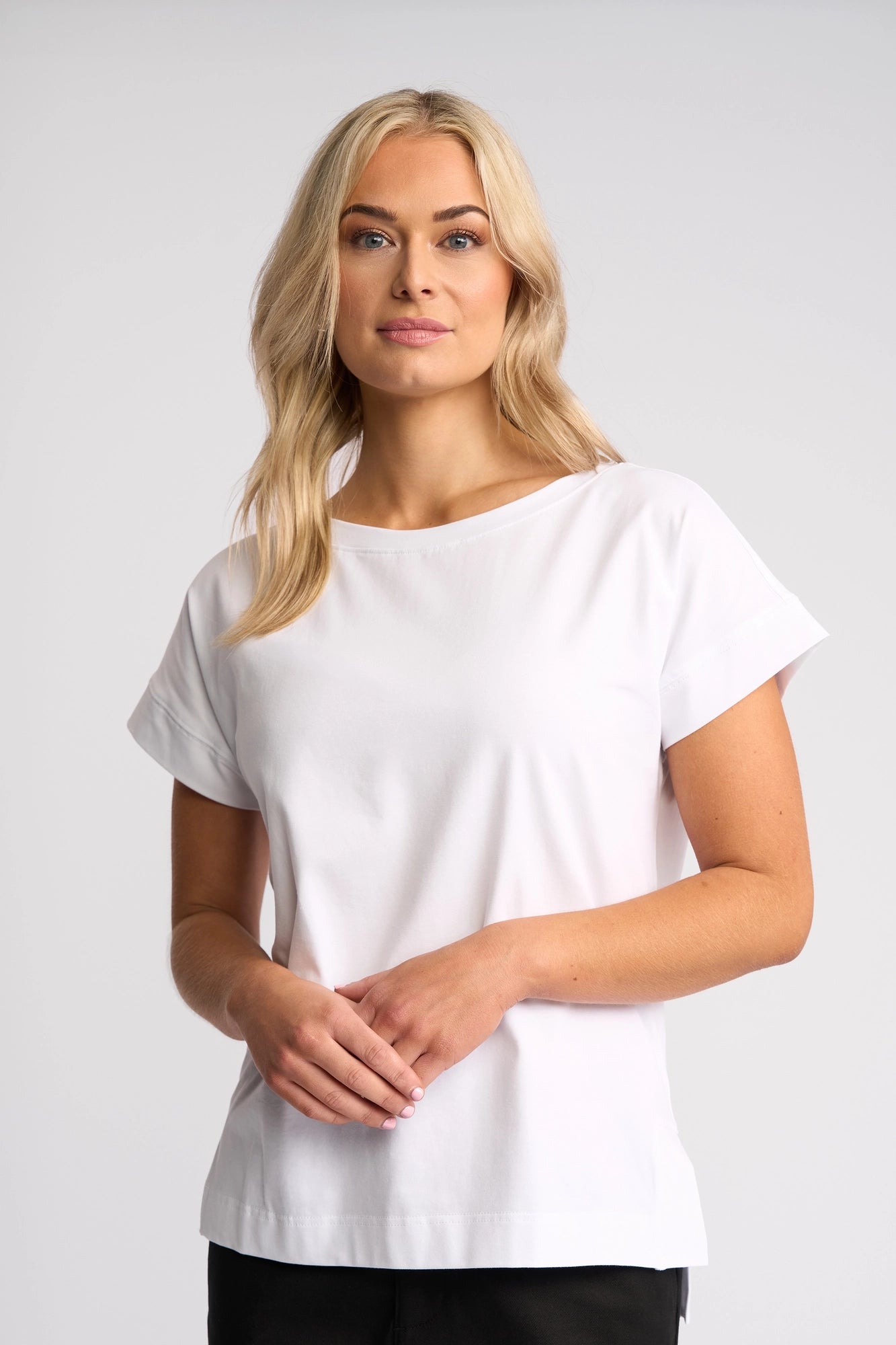 Wearing the Vassalli Thea Top in white, a woman with long blonde hair stands against a light gray background, gazing slightly to the side with a relaxed expression.