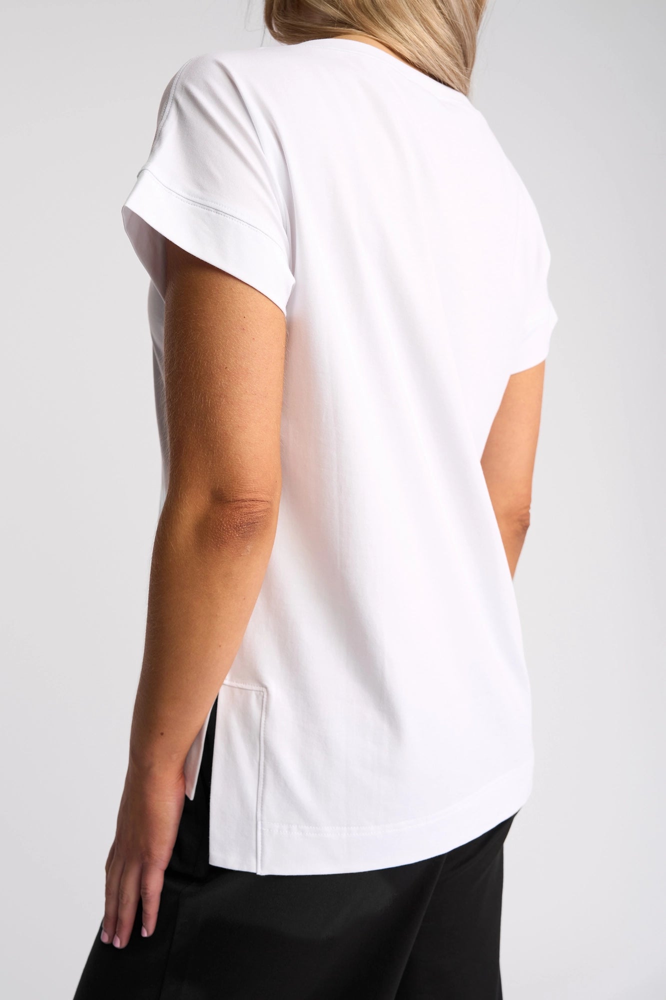 A person with light hair is shown from behind, wearing the Vassalli Thea Top in white—a women’s relaxed t-shirt—paired with black pants, standing against a plain light background.