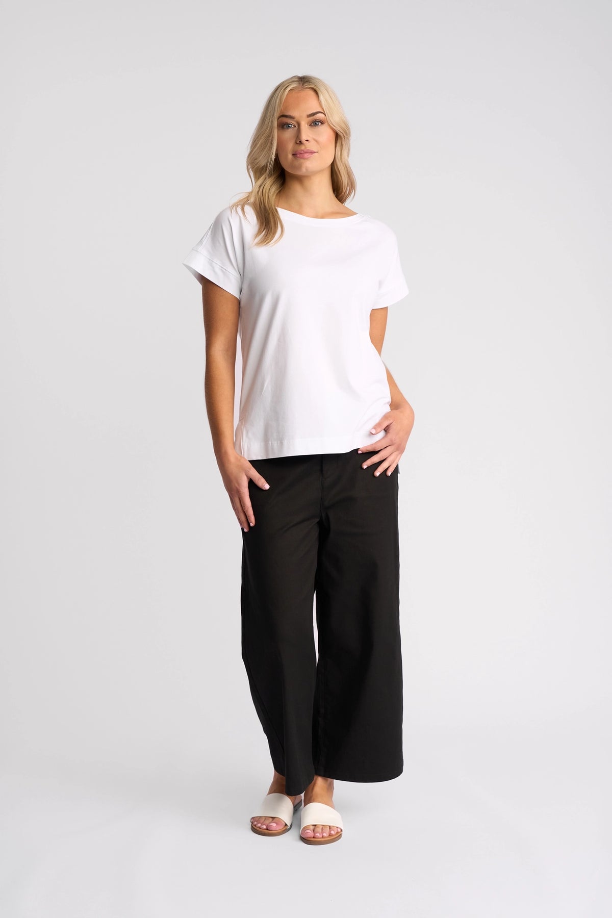 A person with long blonde hair stands against a plain white background, wearing the Vassalli Thea Top in white, loose black pants, and white sandals.