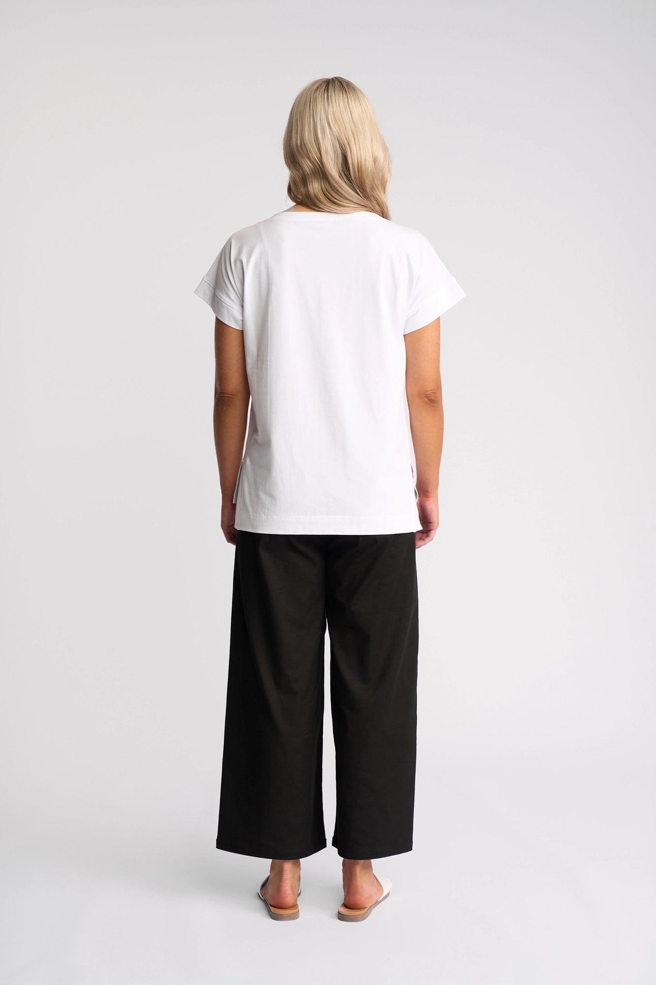 A person with blonde hair stands facing away, wearing the Vassalli Thea Top in white—a women’s relaxed-fit tee—paired with loose black pants and sandals against a plain light background.