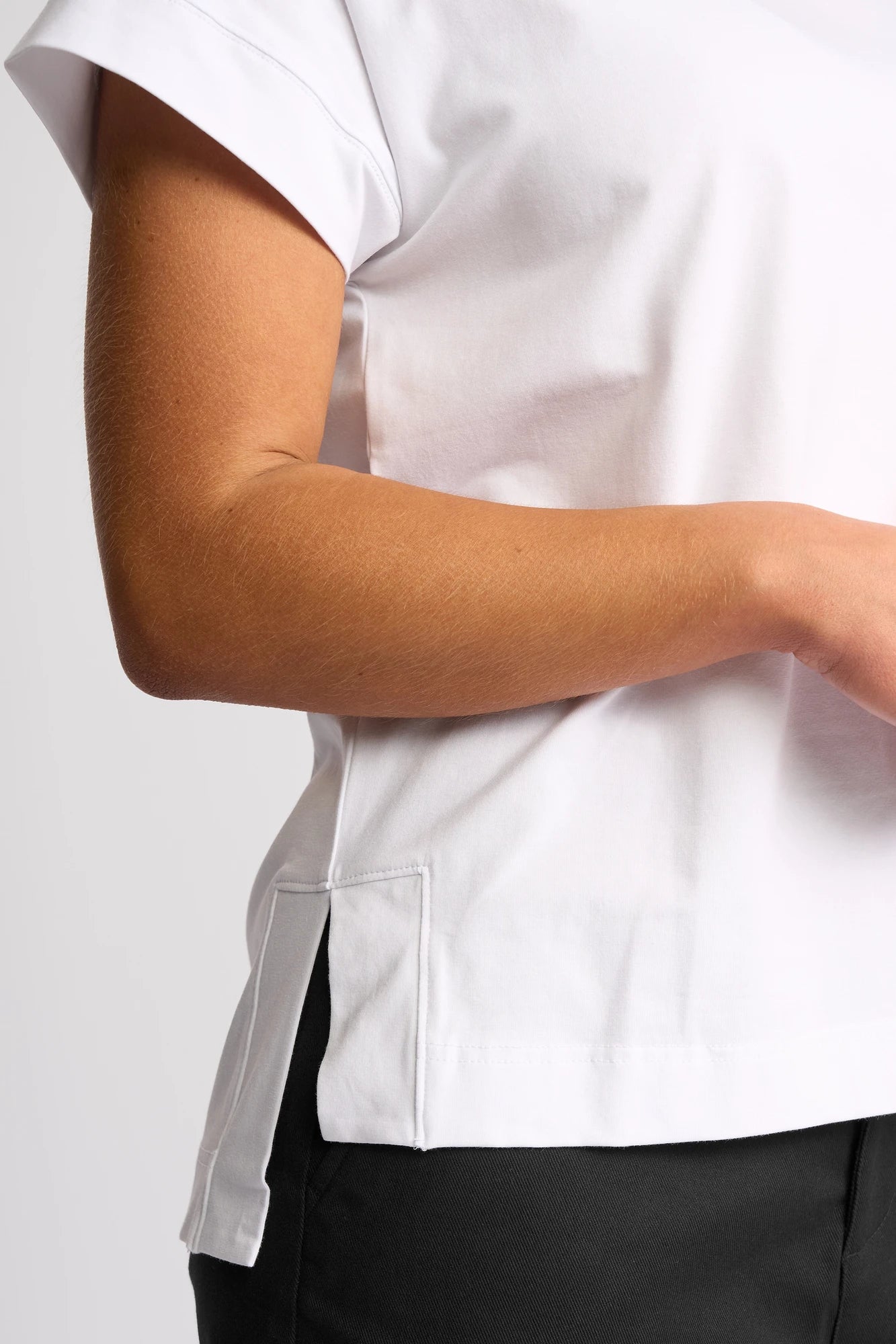 A person is wearing the Vassalli Thea Top - White, a women’s relaxed short-sleeve cotton shirt, paired with black pants. They are standing with one arm bent, revealing the side slit of the Vassalli top and part of their torso.