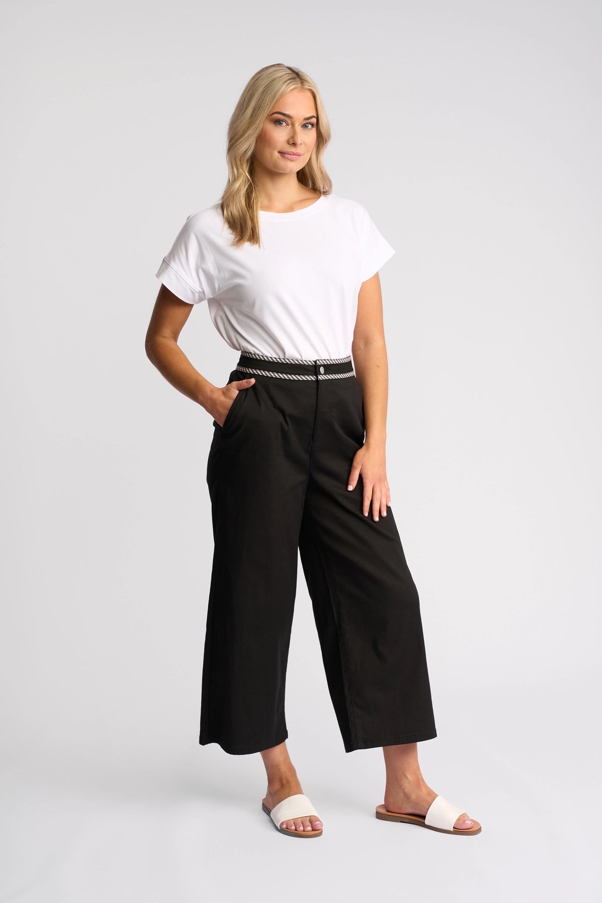 A blonde-haired woman stands against a white background wearing the Vassalli Thea Top in White—an effortlessly stylish, relaxed-fit women’s t-shirt. She pairs it with black wide-leg pants featuring a braided belt and white slide sandals, her hand tucked casually in her pocket and her expression relaxed.