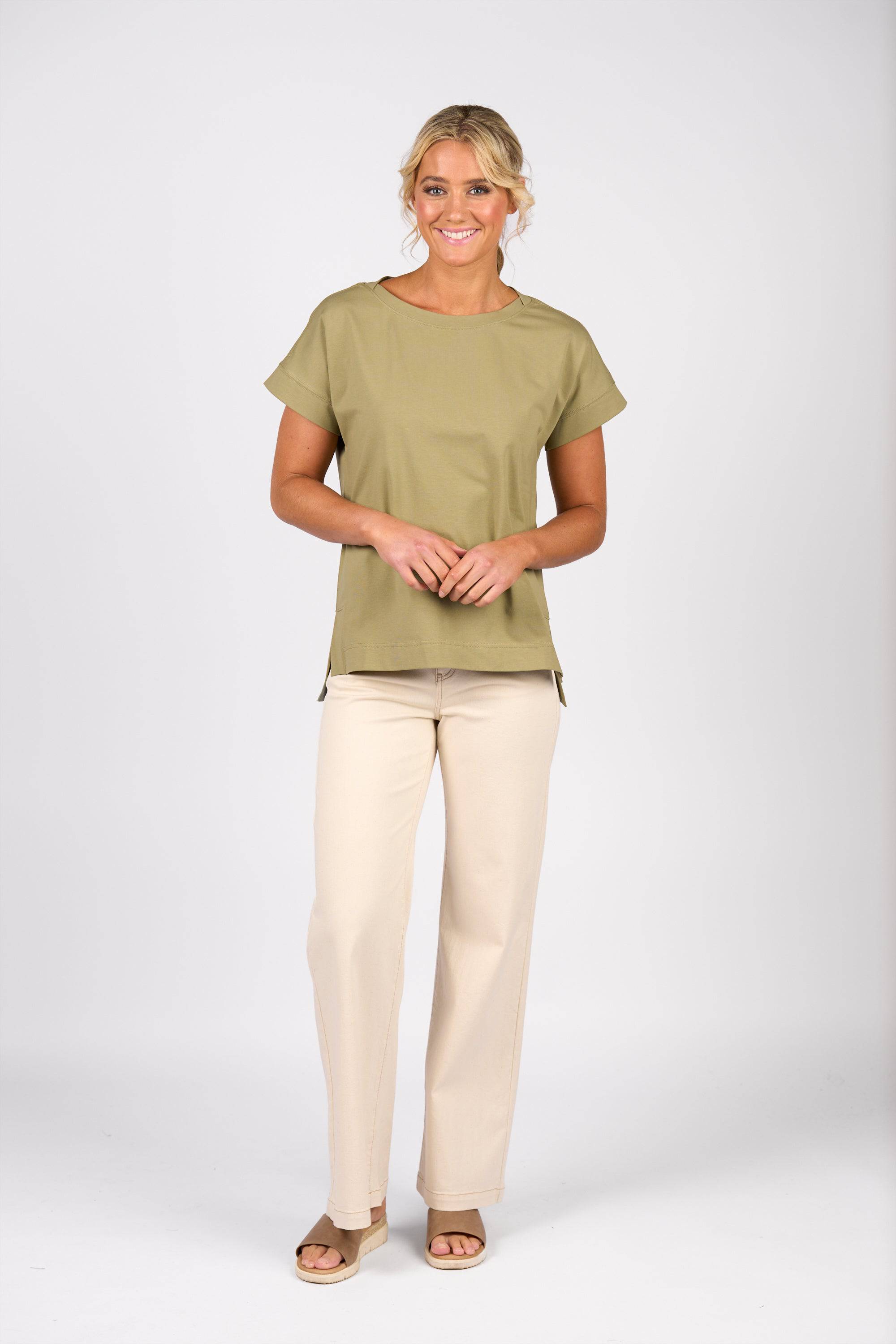 A woman with blonde hair in a loose updo smiles against a plain white background, wearing the Vassalli Thea Top in sage, beige pants, and tan sandals.