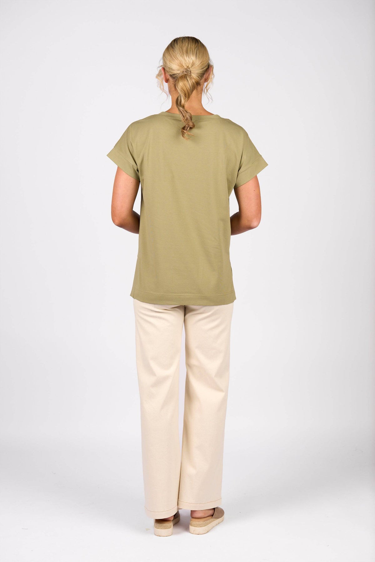 A person with blonde hair in a low braid is seen from behind wearing the Vassalli Thea Top in sage, paired with cream pants and beige shoes against a plain white background.