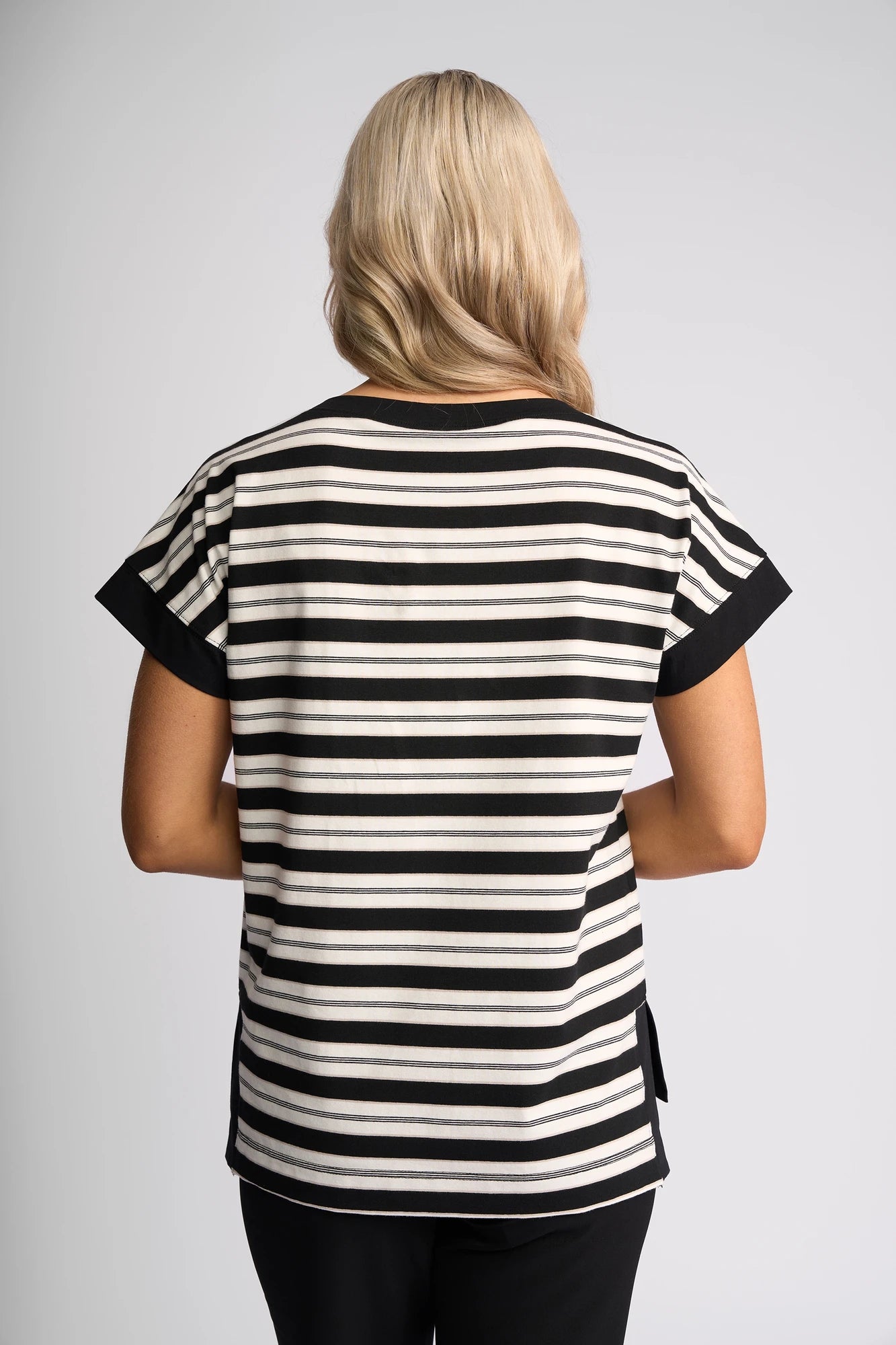 A person with long blonde hair stands facing away, wearing the Vassalli Thea Top in black and white stripes paired with black pants, set against a plain light background.