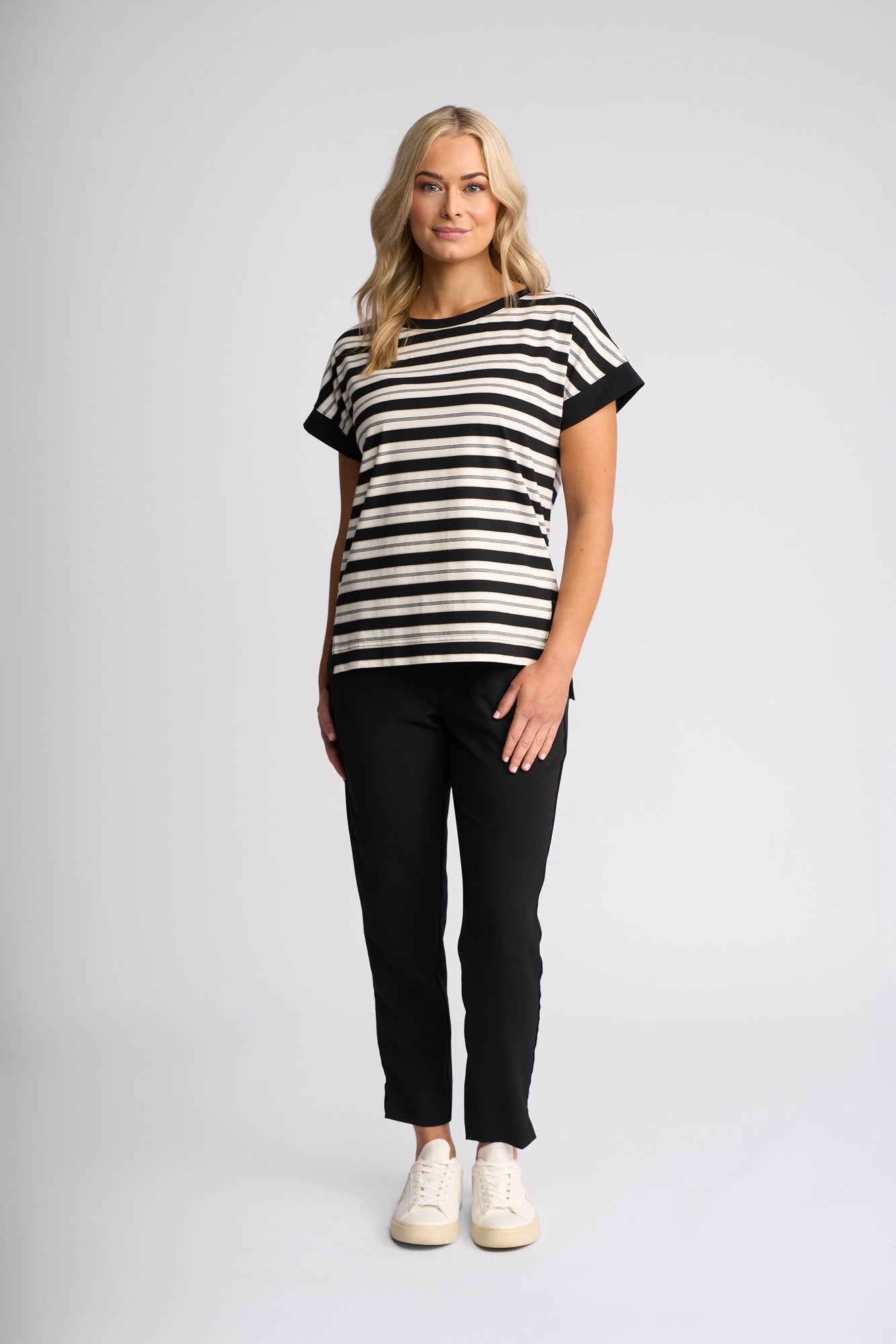 A woman with long blonde hair stands and smiles against a plain light background, wearing the Vassalli Thea Top in Black/White, paired with black pants and white sneakers.