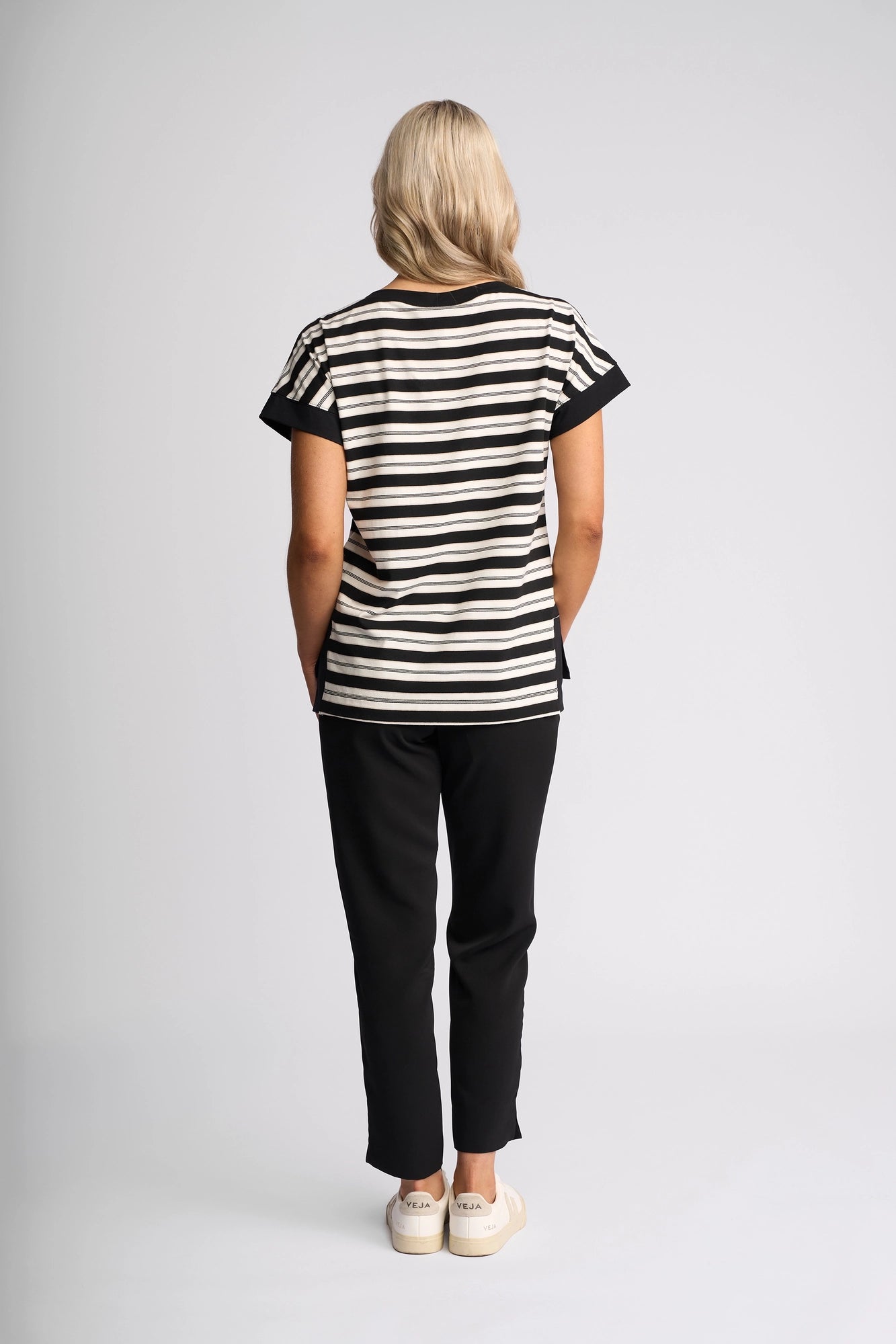 A person with long blonde hair stands facing away, wearing the Vassalli Thea Top - Black/White styled as a women’s boat neck shirt, paired with black pants and white sneakers against a plain light background.