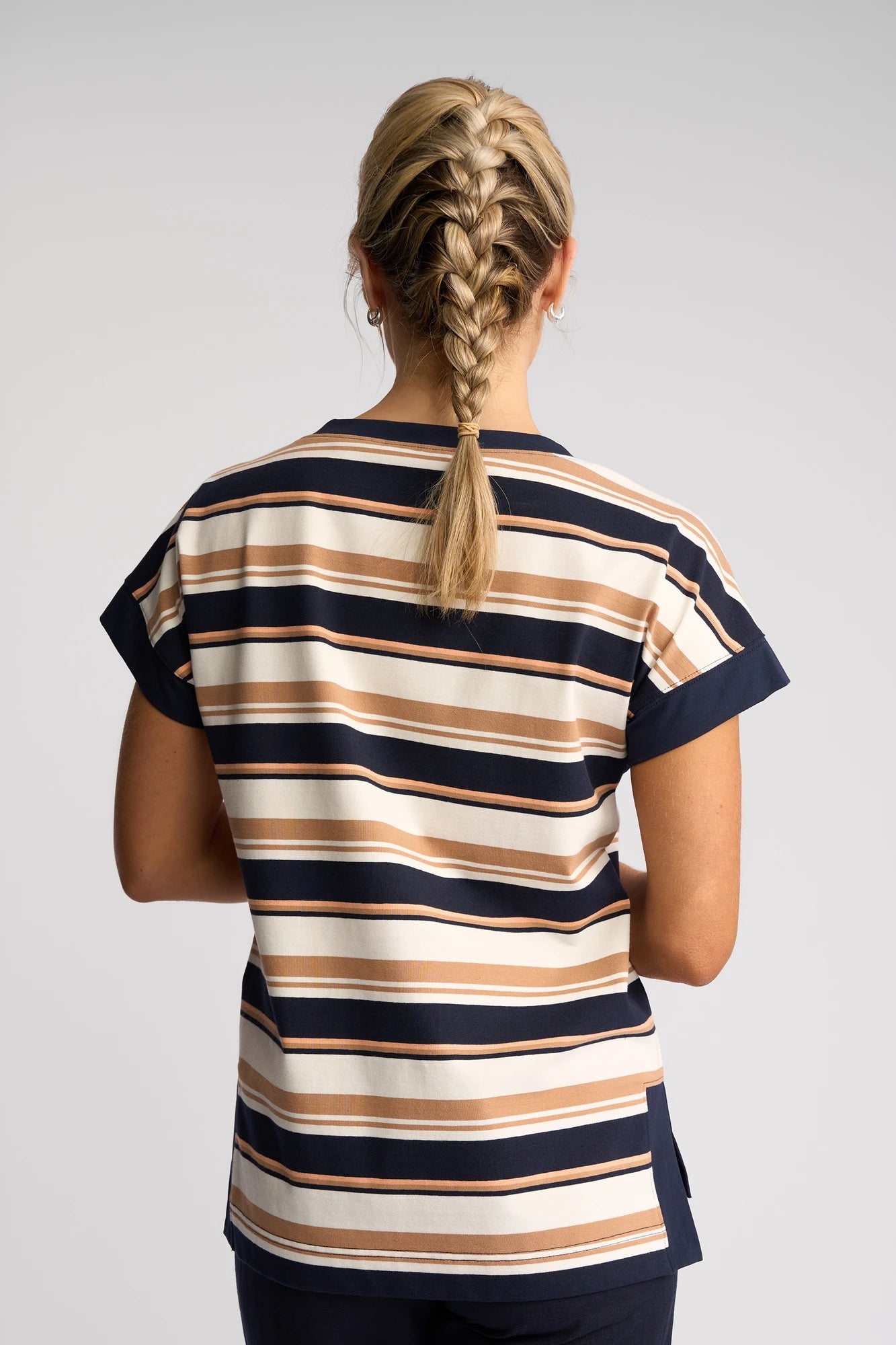 A person with blonde braided hair is shown from behind, wearing the Vassalli Thea Top - Multi Stripe— a short-sleeve cotton top featuring navy, beige, and cream stripes with a stylish boat neckline—set against a simple light background.