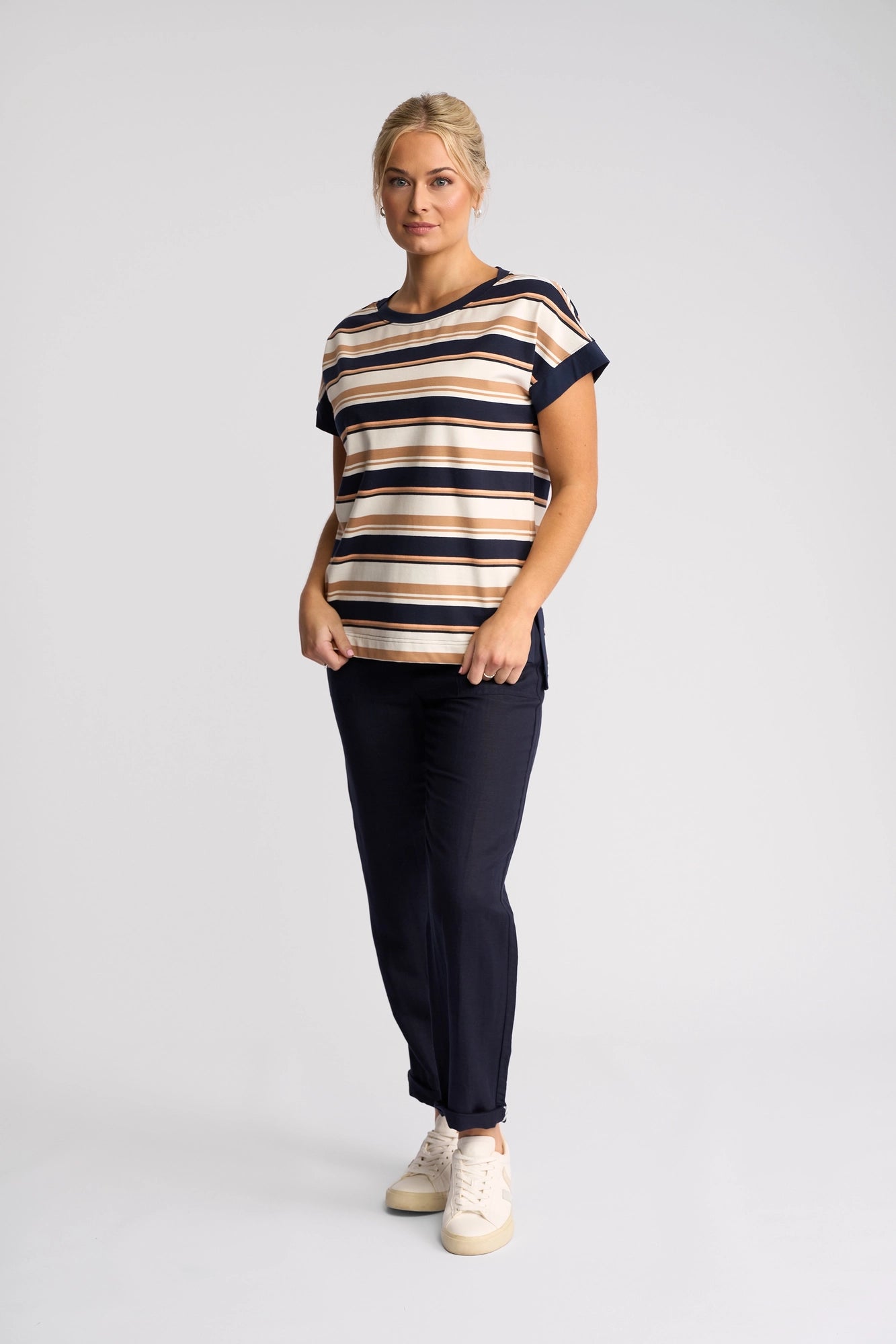 A woman with blond hair tied back stands against a plain background, wearing the Vassalli Thea Top - Multi Stripe, featuring short sleeves, a boat neckline, and neutral-toned stripes. She pairs it with dark pants and white sneakers, looking slightly to the side with a neutral expression.
