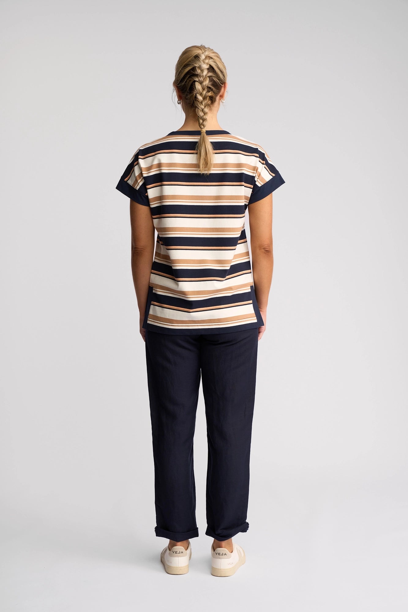 A person with blonde braided hair stands facing away, wearing the Vassalli Thea Top – Multi Stripe, a short-sleeve cotton top featuring a boat neckline and multi-colored stripes. They pair it with dark pants and light-colored sneakers against a simple, light background.