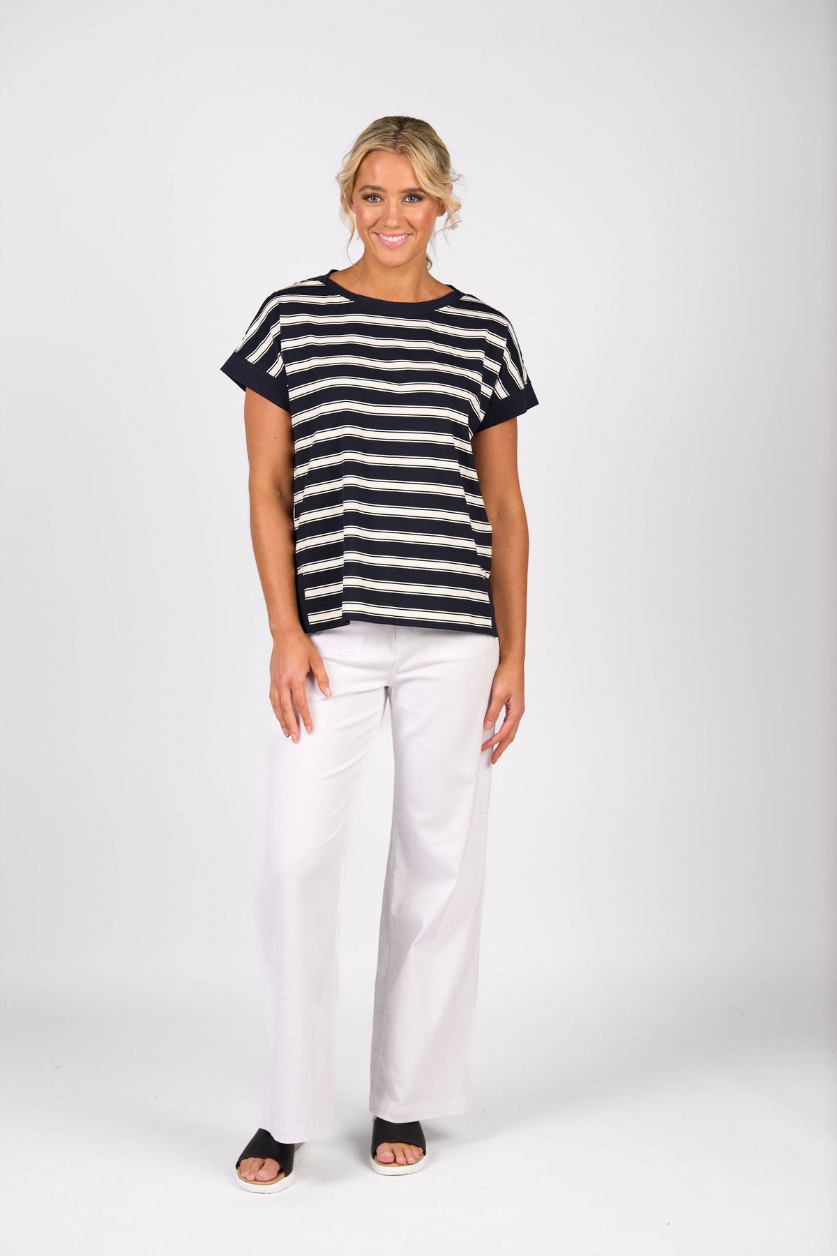 A woman smiles against a white background, wearing the Vassalli Thea Top in Ink Stripe with short sleeves and a boat-neck, paired with white wide-leg pants and black open-toe sandals. Her short, wavy blonde hair completes the look.