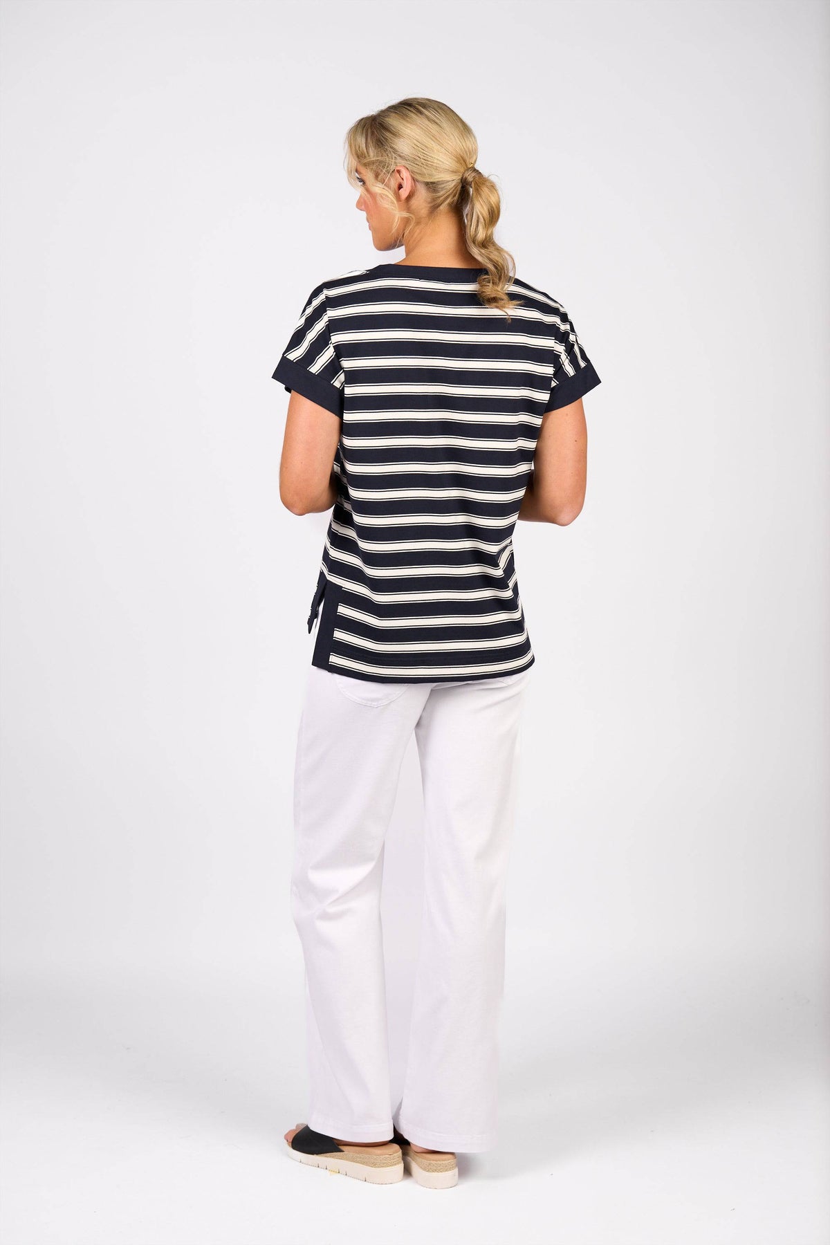 A woman with blonde hair in a low ponytail stands facing away, wearing the Vassalli Thea Top - Ink Stripe, white pants, and black slip-on shoes against a plain white background.