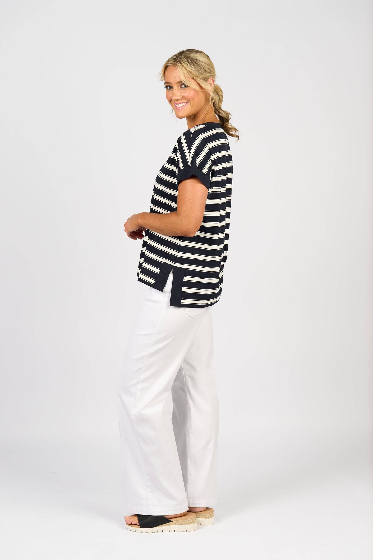 A woman stands sideways, smiling, wearing the Vassalli Thea Top in Ink Stripe with white wide-leg pants and black platform sandals against a plain white background.