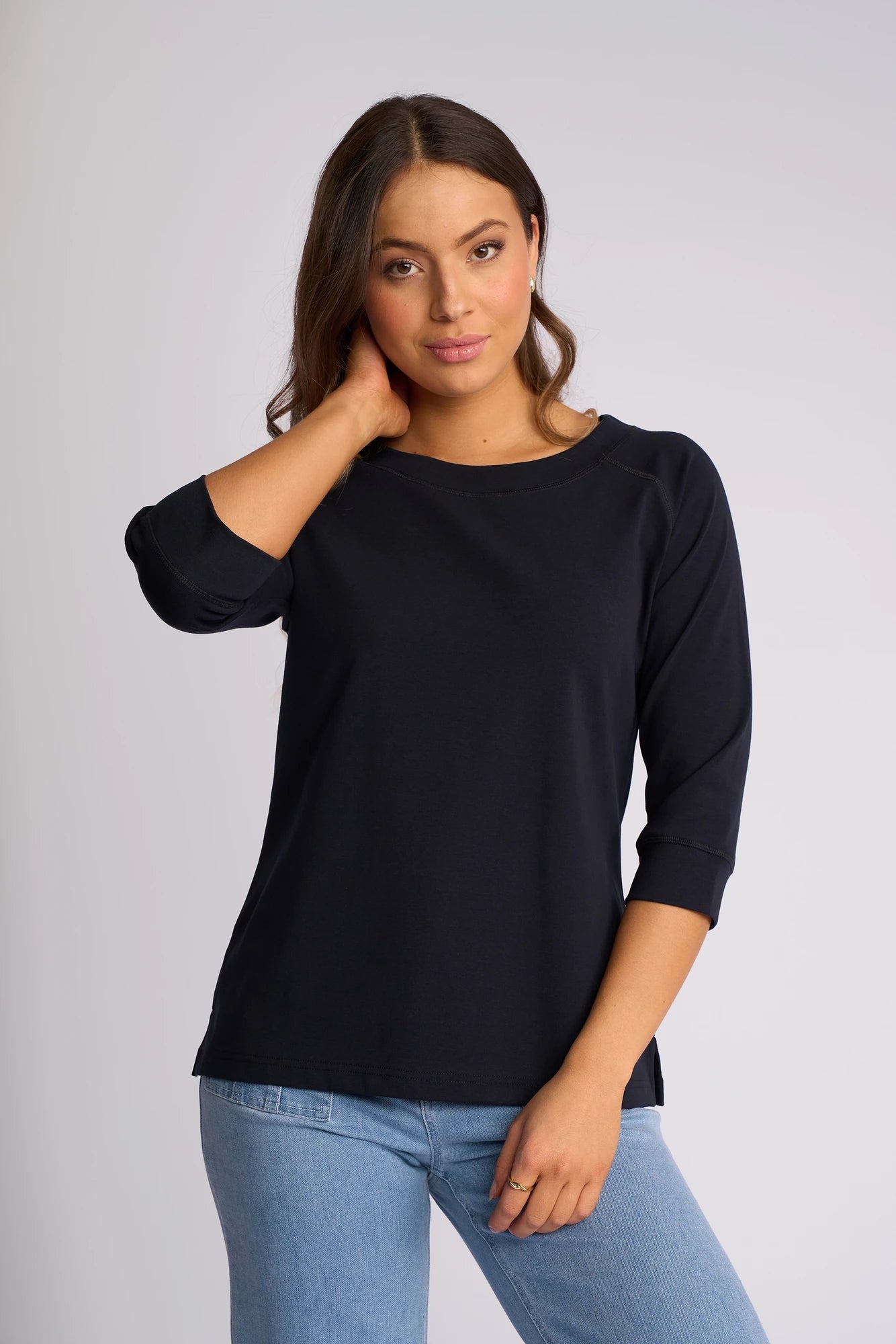 A woman with long brown hair stands against a plain light background, touching her neck with one hand and looking at the camera. She is wearing a Vassalli Canyon Top - Ink with 3/4 length sleeves and light blue jeans. The top is made from a comfortable cotton-elastane blend.